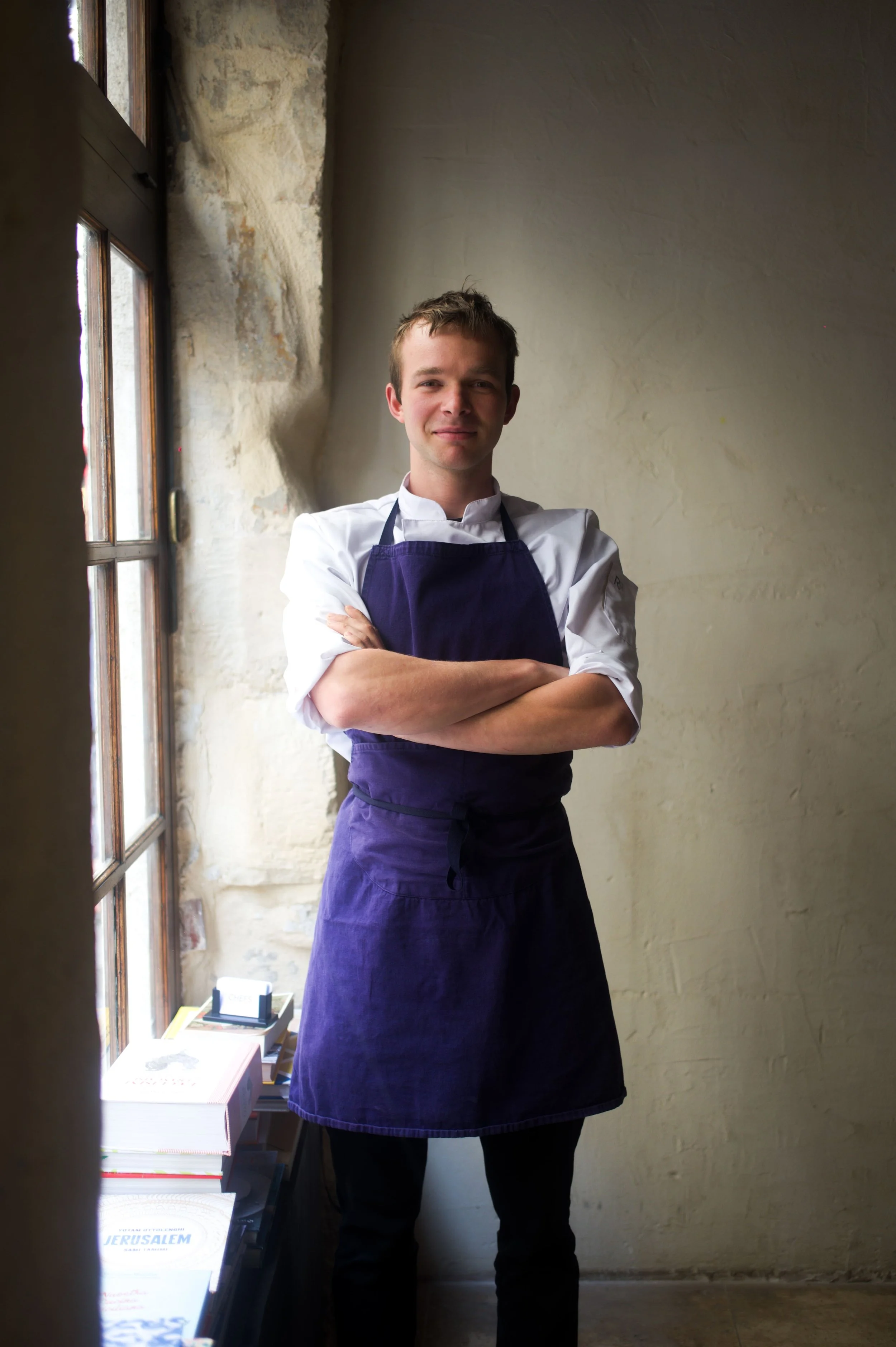 Jeune chef en uniforme blanc et tablier violet, debout avec les bras croisés près d'une fenêtre dans une cuisine ou un restaurant, souriant.