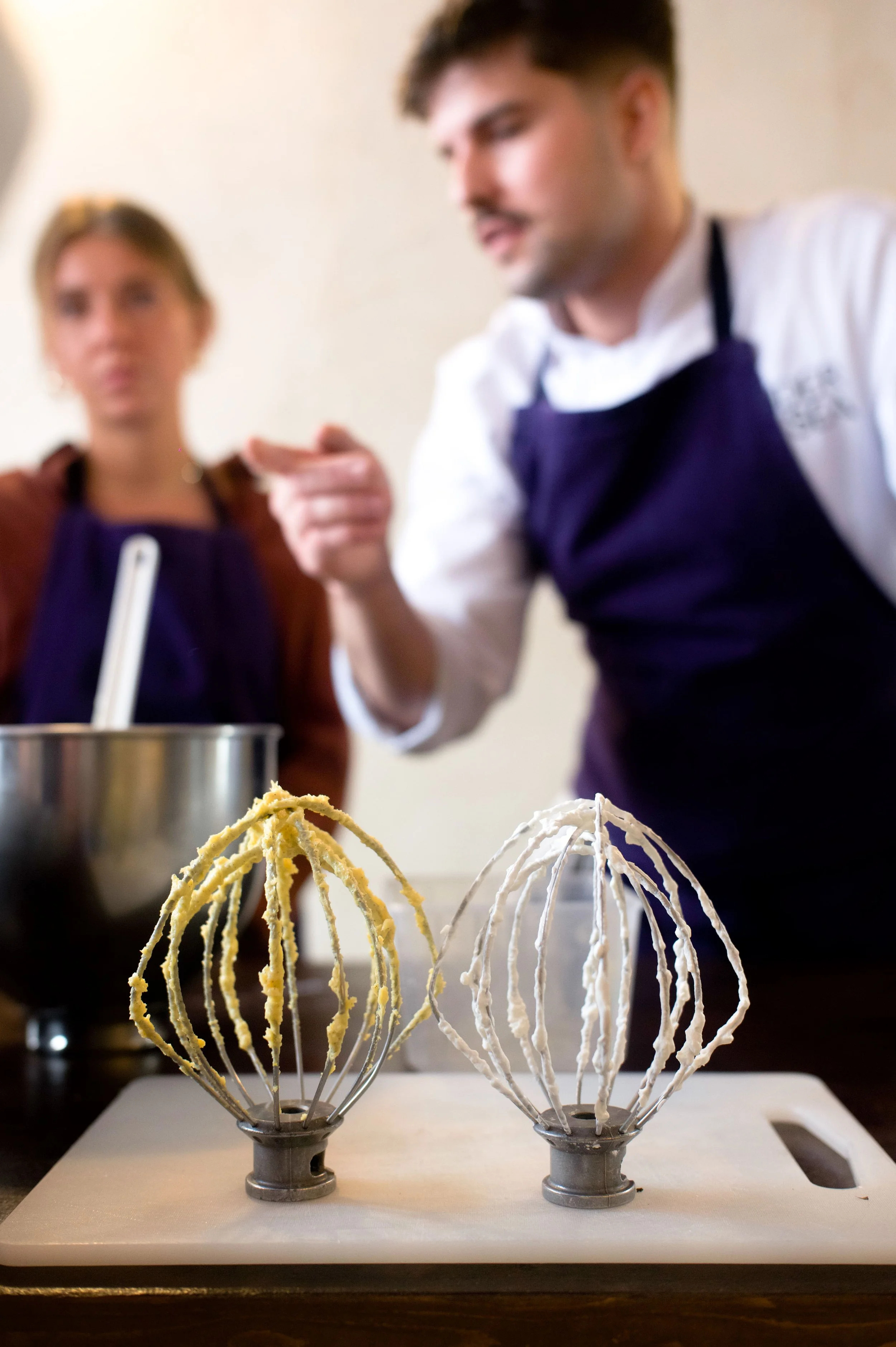 Deux fouets avec de la pâte à gâteau, un à gauche avec de la pâte jaune et un à droite avec de la pâte blanche, sur une planche en bois, avec deux personnes en arrière-plan parlant devant un mur beige, une femme et un homme portant un tablier violet