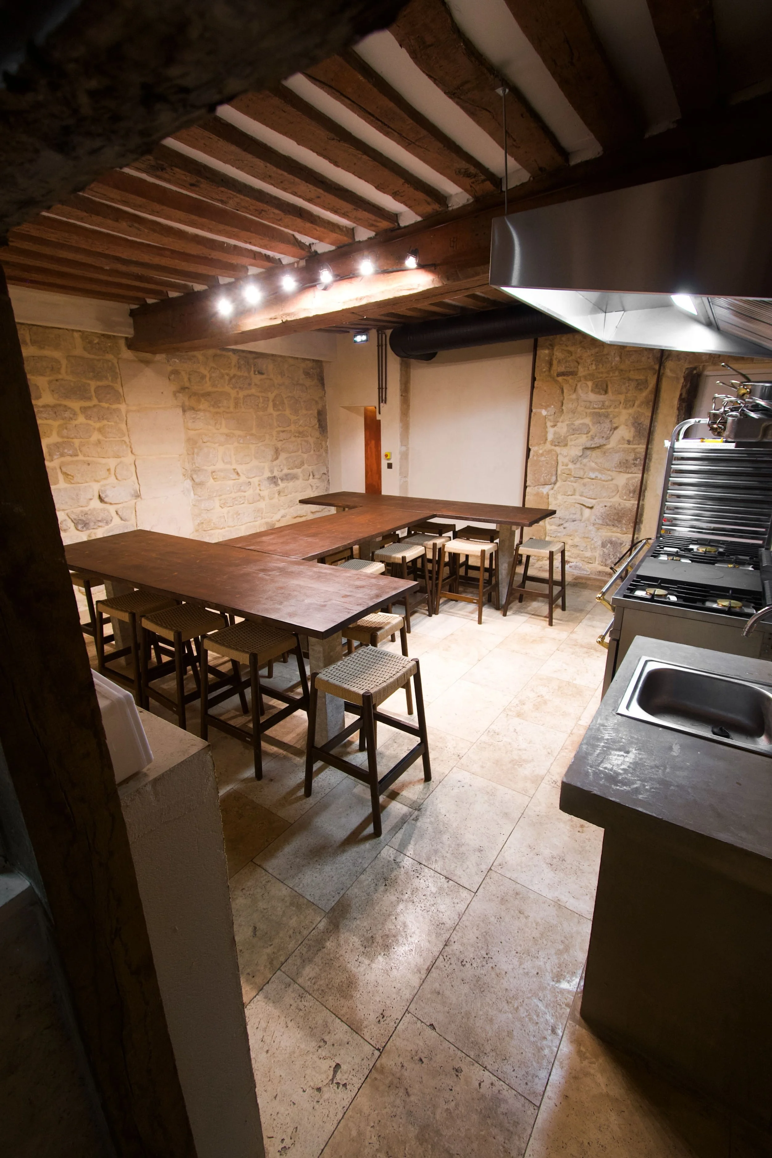 A kitchen or dining area with wooden tables, chairs and a stone wall, lit by a hanging light, in a rustic style.