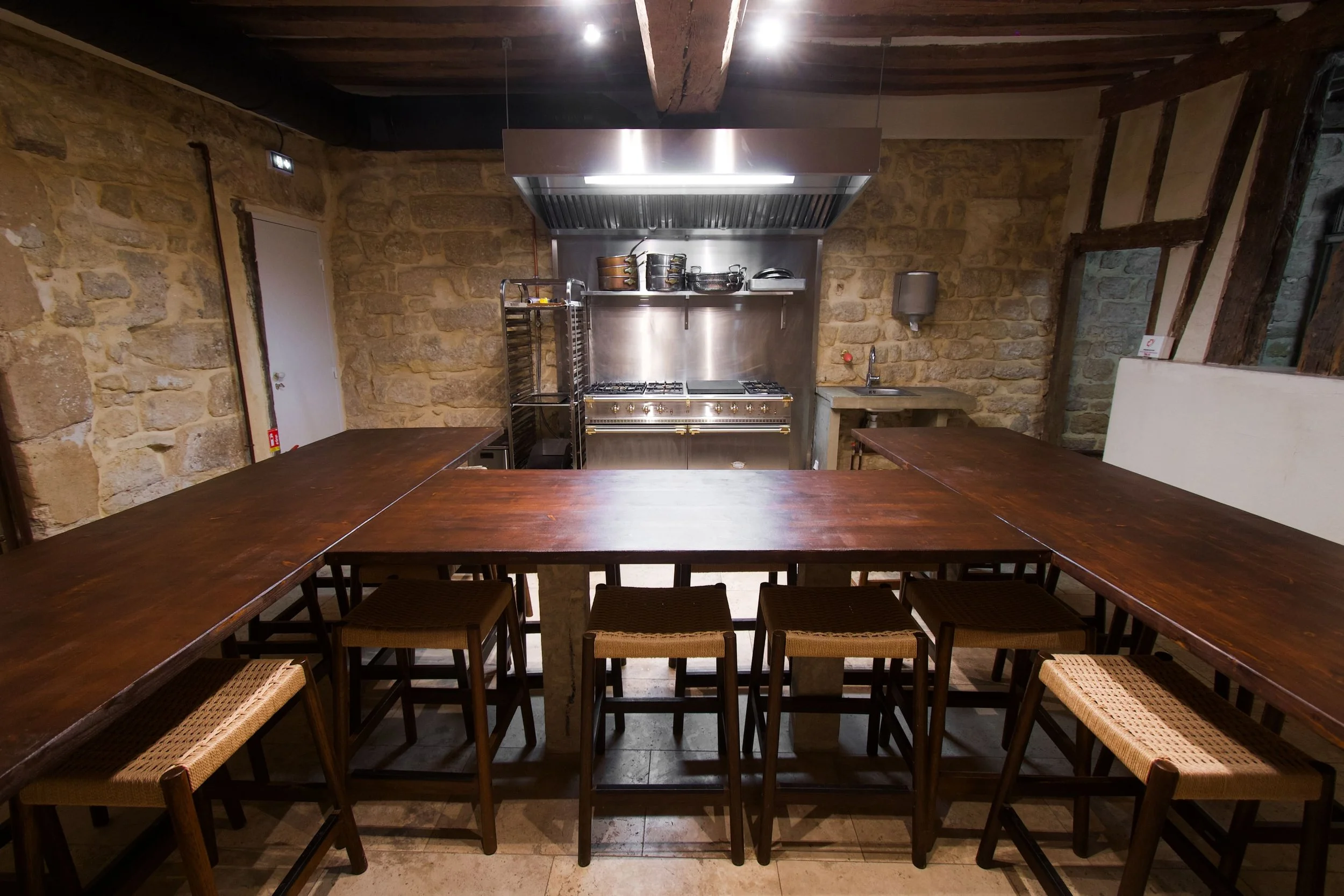 Rustic kitchen with large wooden table, wooden chairs with bristle seats, stone wall, stainless steel kitchen with stove, shelves with cooking utensils, sink and a few tools.
