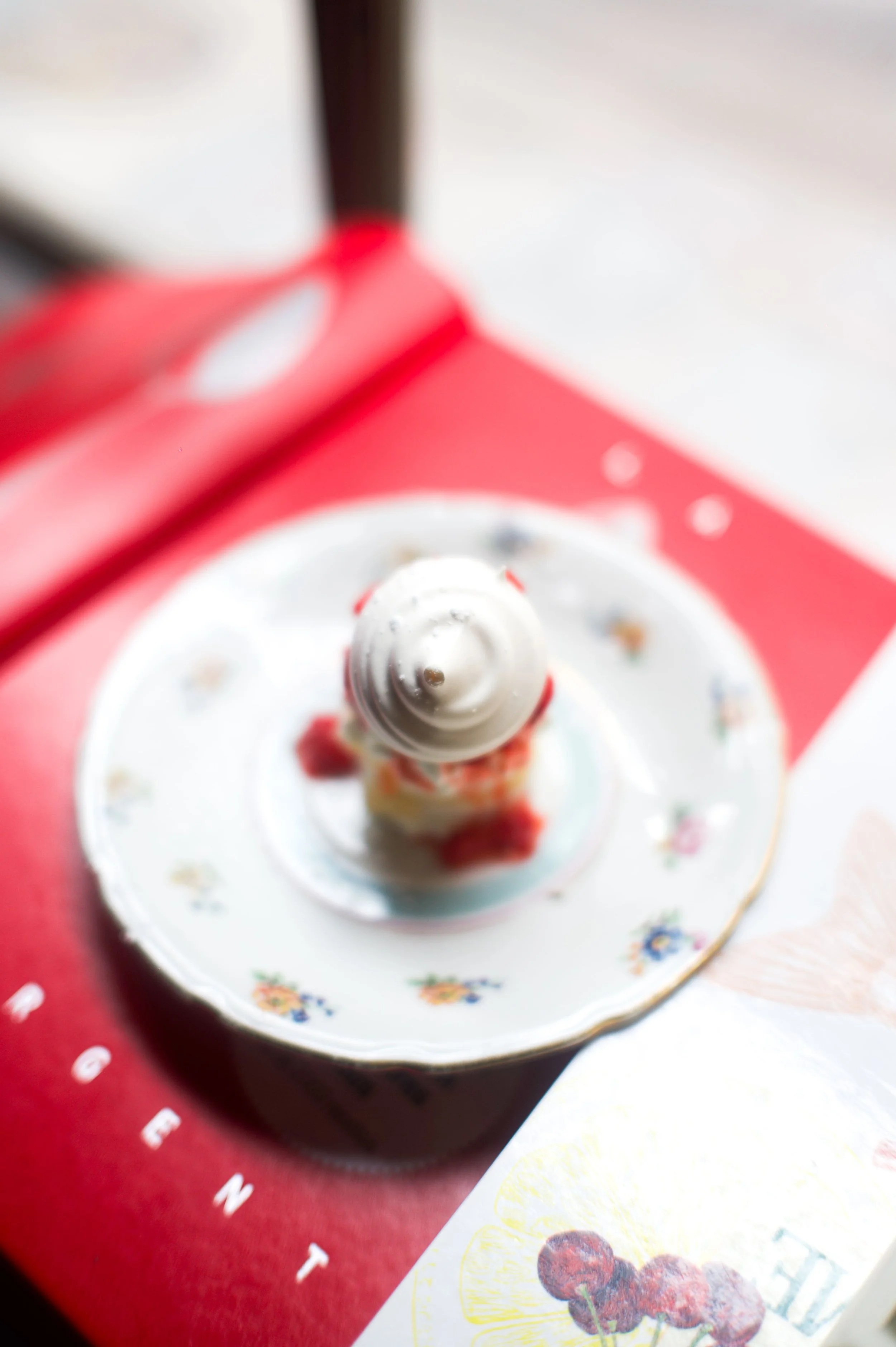Photo d'une glace en forme de cône, décorée avec des fruits, placée sur une assiette en porcelaine avec motifs floraux, sur fond rouge avec le mot 'COGNAC' visible.