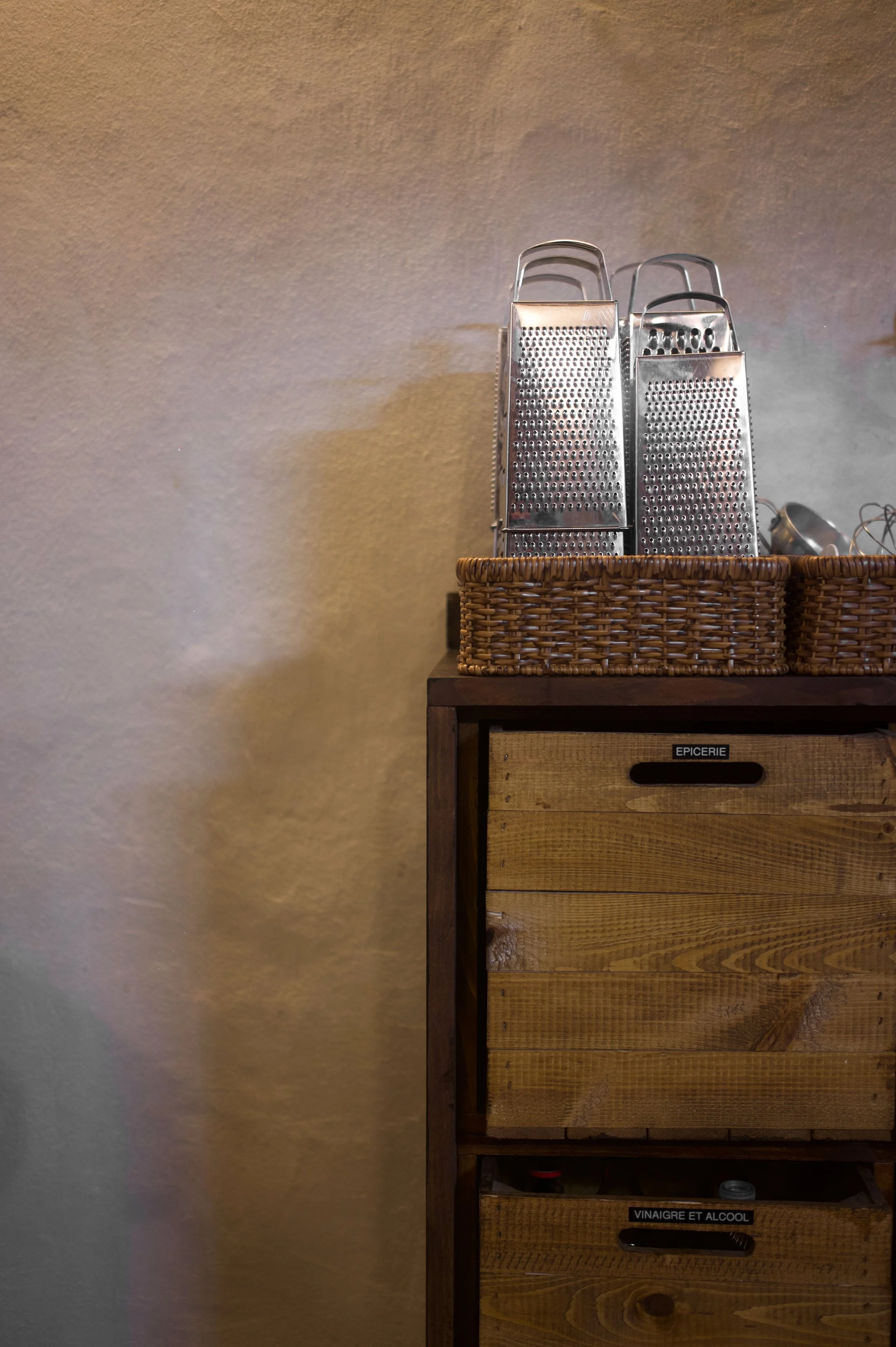 Une étagère en bois avec deux tiroirs en dessous marqués 'EPICERIE' et 'VINAIGRE ET ALCOOL', avec des paniers en osier contenant des râpes à fromage en métal sur le dessus contre un mur beige.