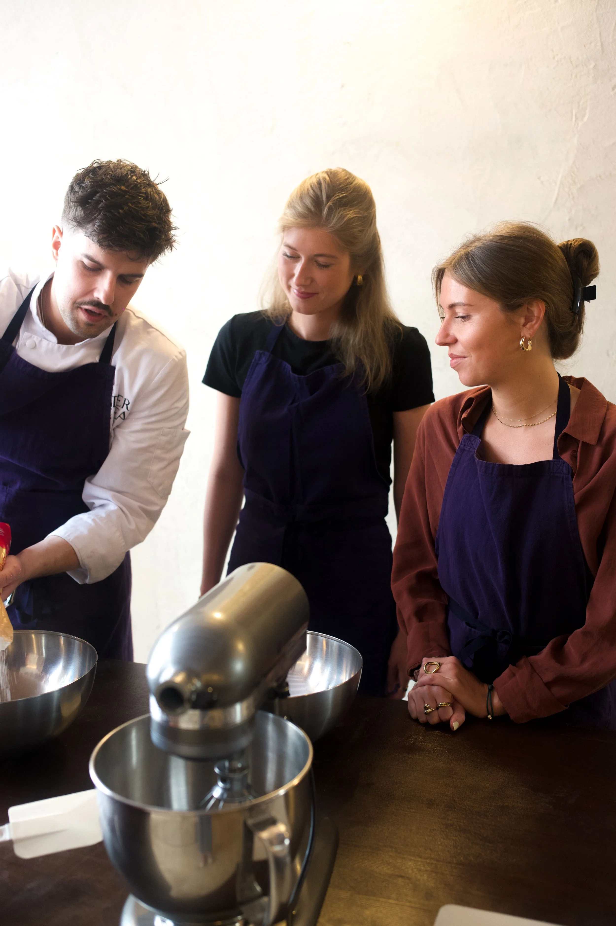 Three people looking at an electric mixer during a cooking workshop, two women and a man all wearing purple aprons.