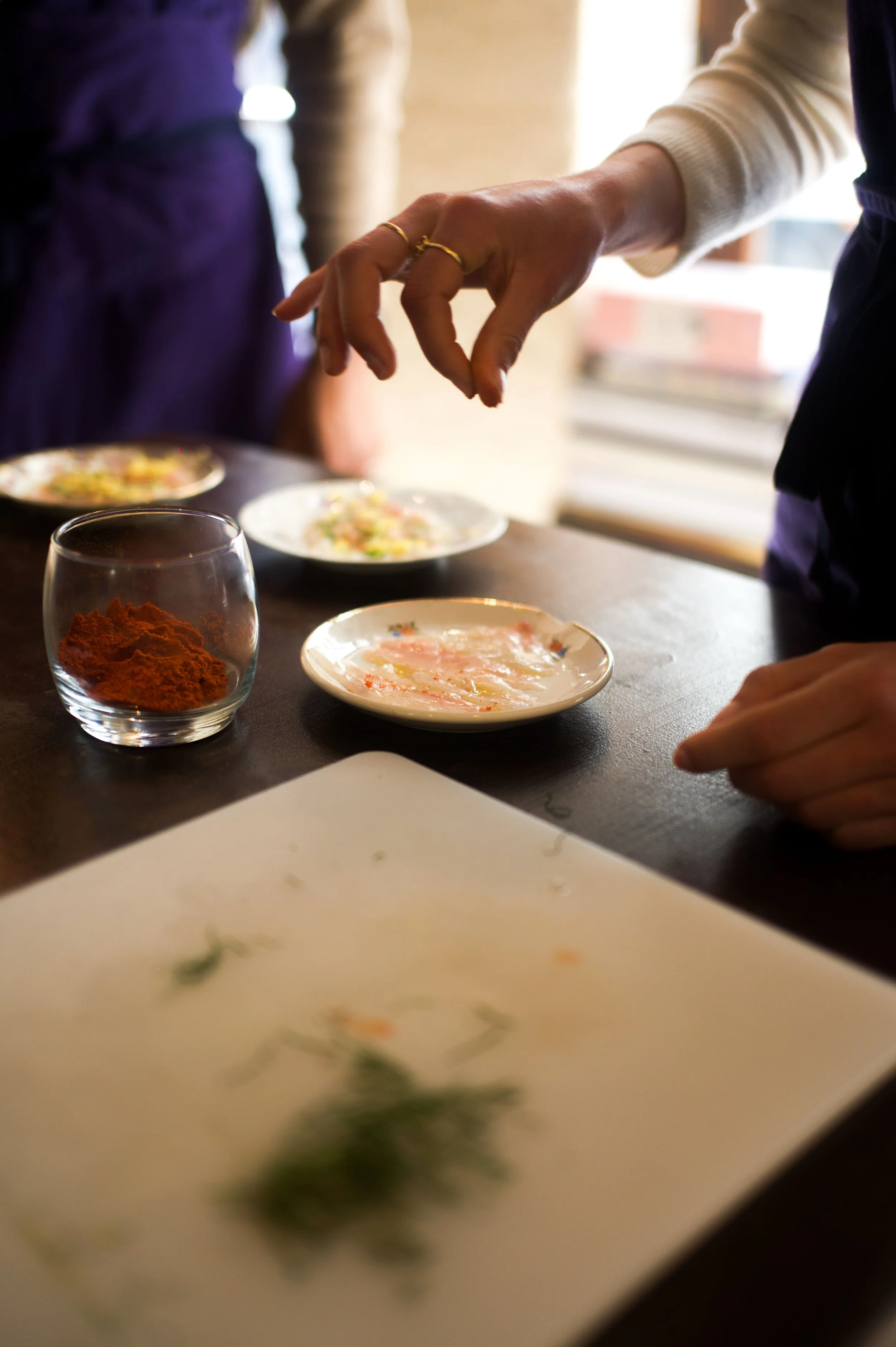 Personne qui prépare des épices et des ingrédients pour une recette dans une cuisine, avec des plats en arrière-plan.