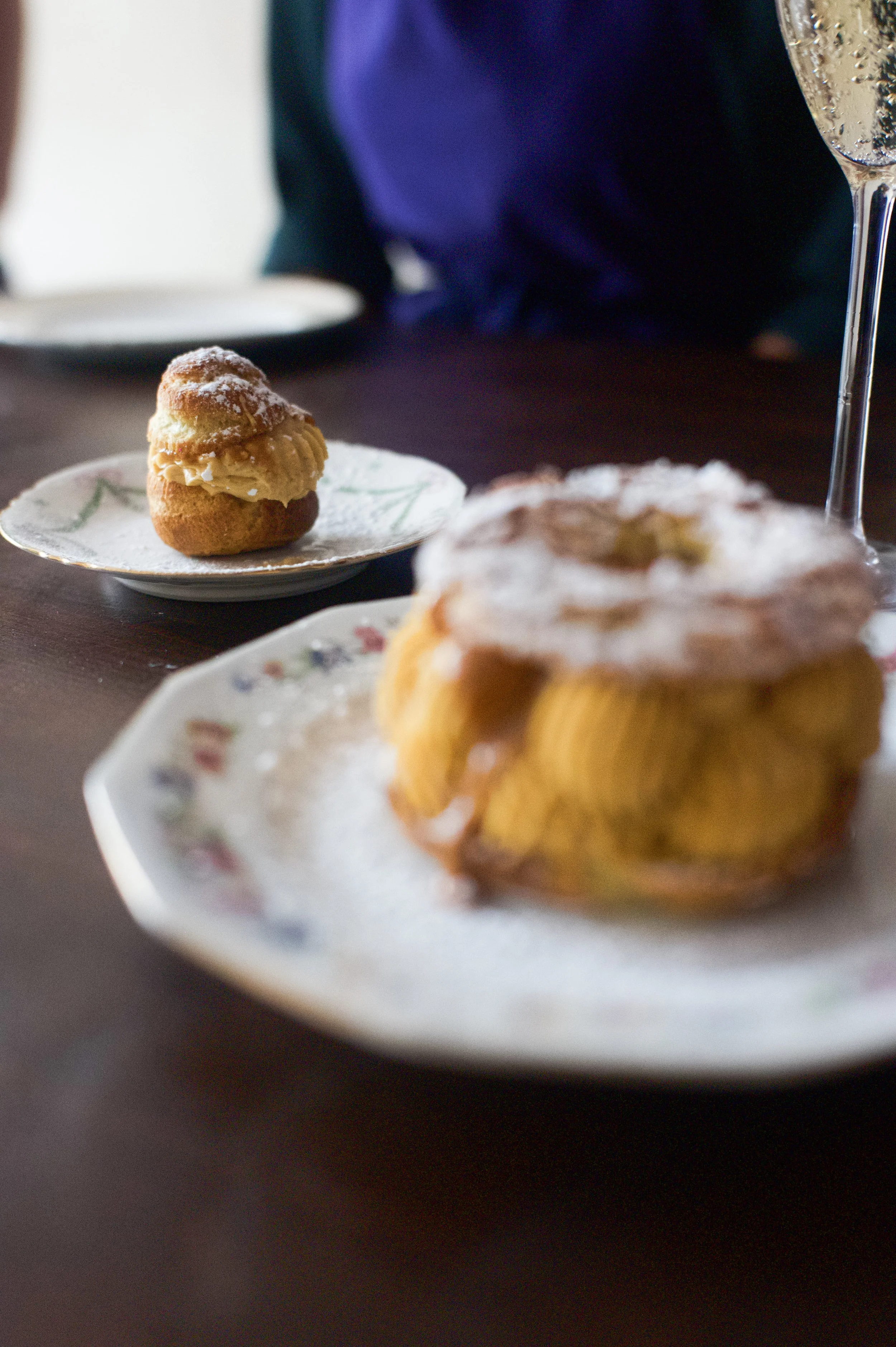 Deux desserts, une religieuse et une tartelette, servis sur des assiettes à motif floral, avec un verre de champagne en arrière-plan.