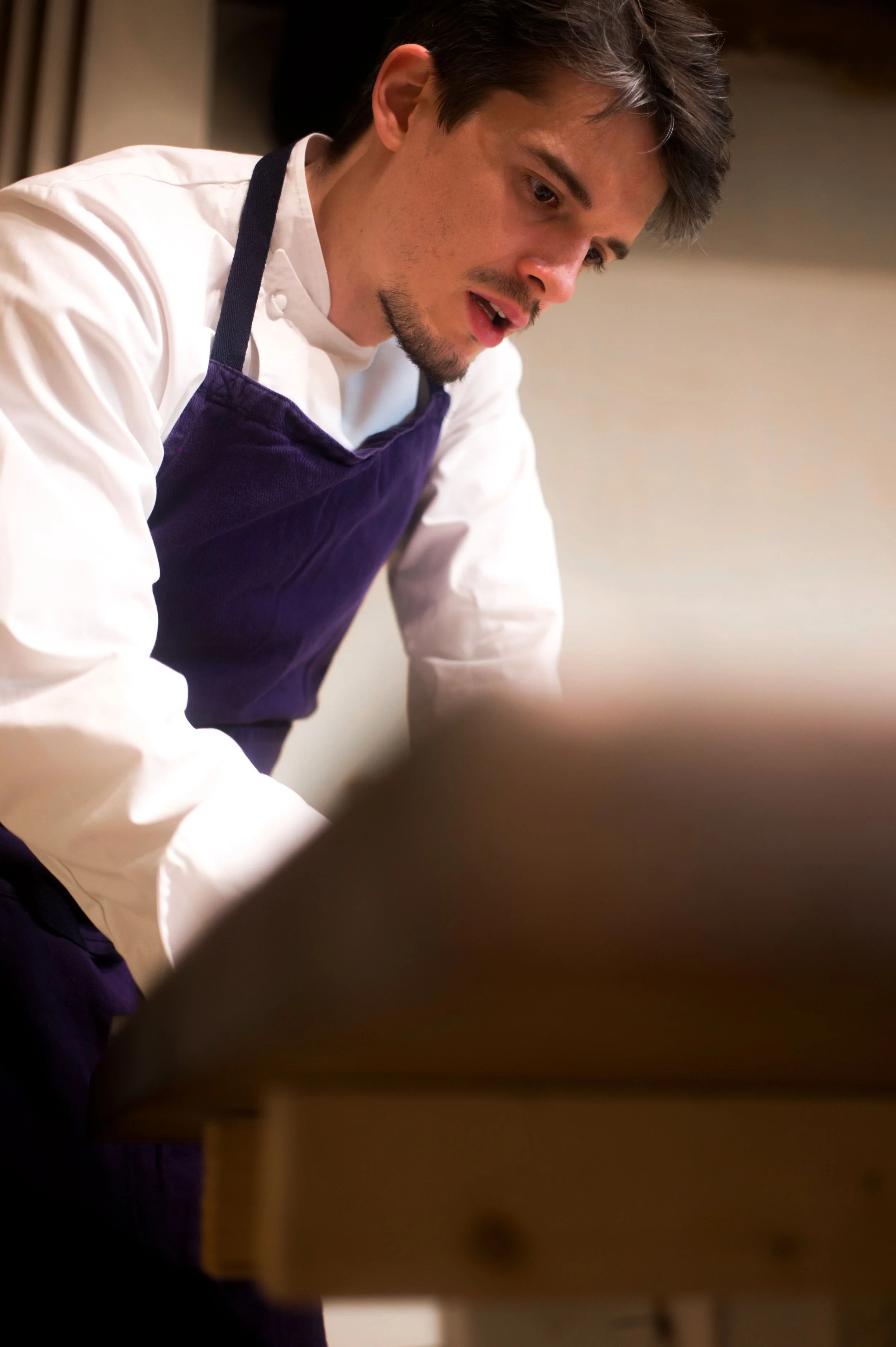 Un homme portant un uniforme de cuisine, une chemise blanche et un tablier violet, en train de cuisiner ou de préparer quelque chose.