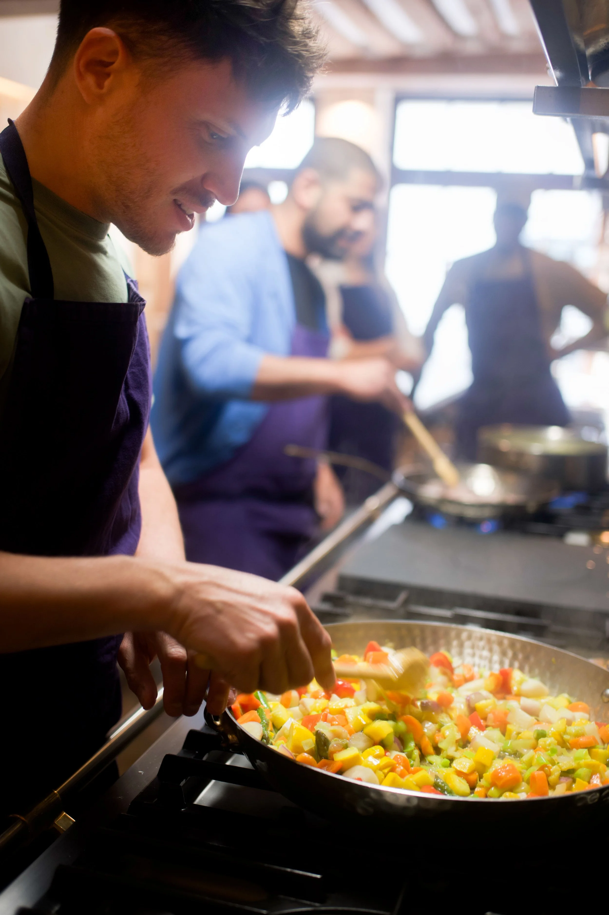 Un jeune homme cuisine des légumes dans une poêle en cuisine professionnelle, avec deux autres personnes en arrière-plan qui travaillent également.