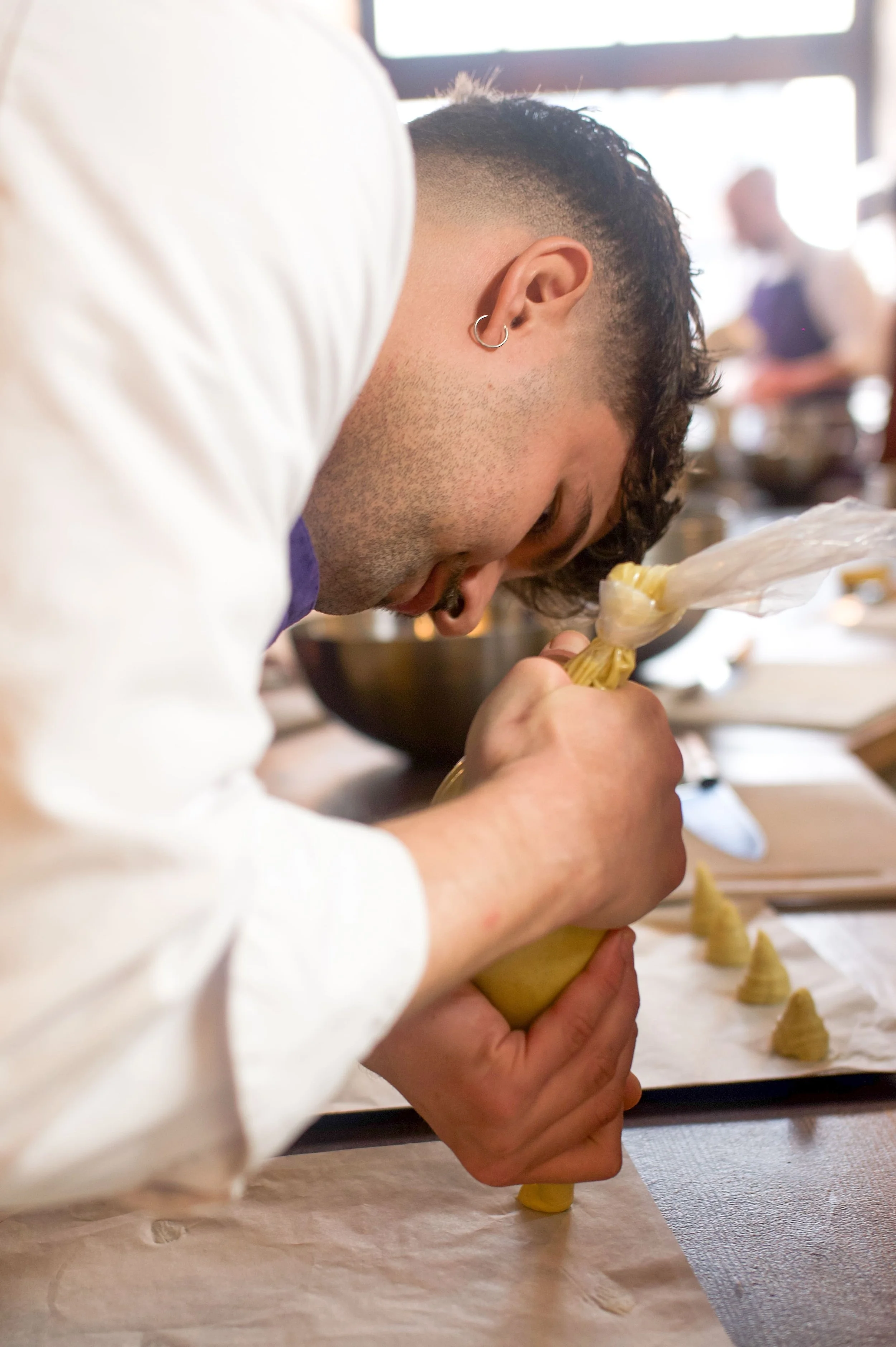 Un jeune homme avec des boucles d'oreilles déposé de côté, en train de faire de la pâte ou de la nourriture avec une poche à douille, dans une cuisine ou un atelier culinaire, avec d'autres personnes en arrière-plan.