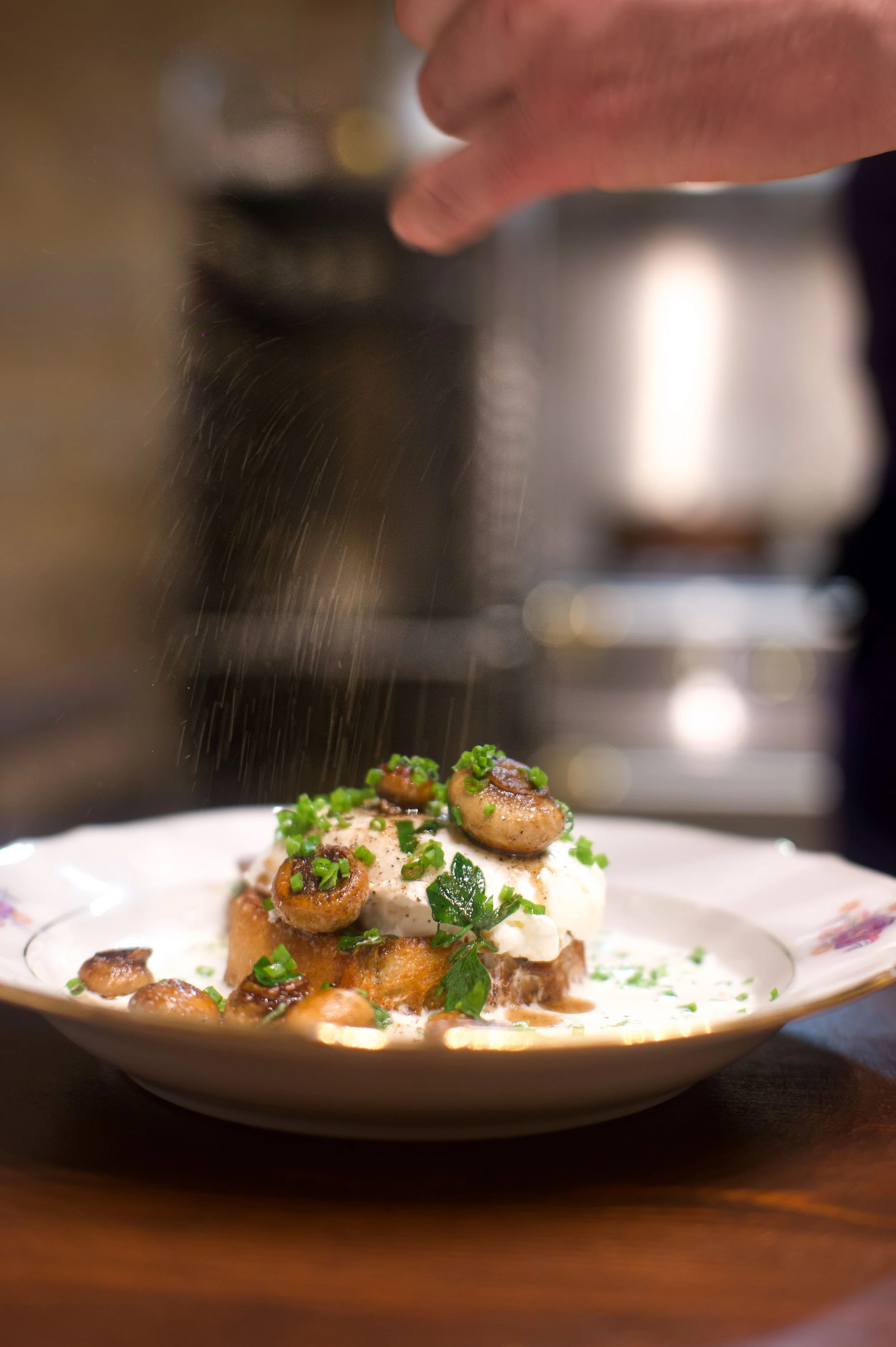 Une assiette de repas avec des champignons, de la crème et des herbes, avec une personne en train de saupoudrer un ingrédient dessus.