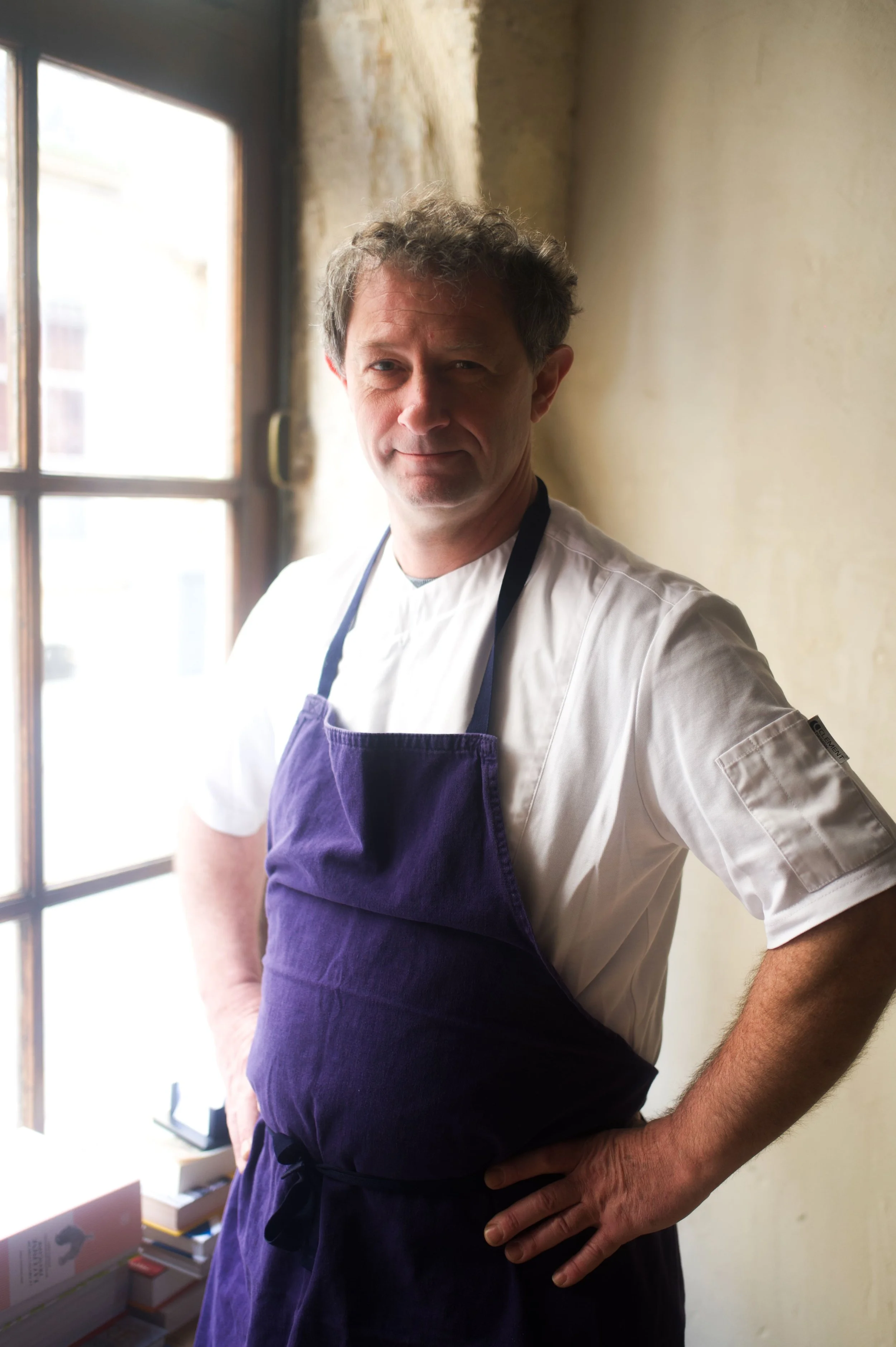 Homme chef cuisinier en uniforme blanc et tablier violet, posant à côté d'une fenêtre dans une cuisine
