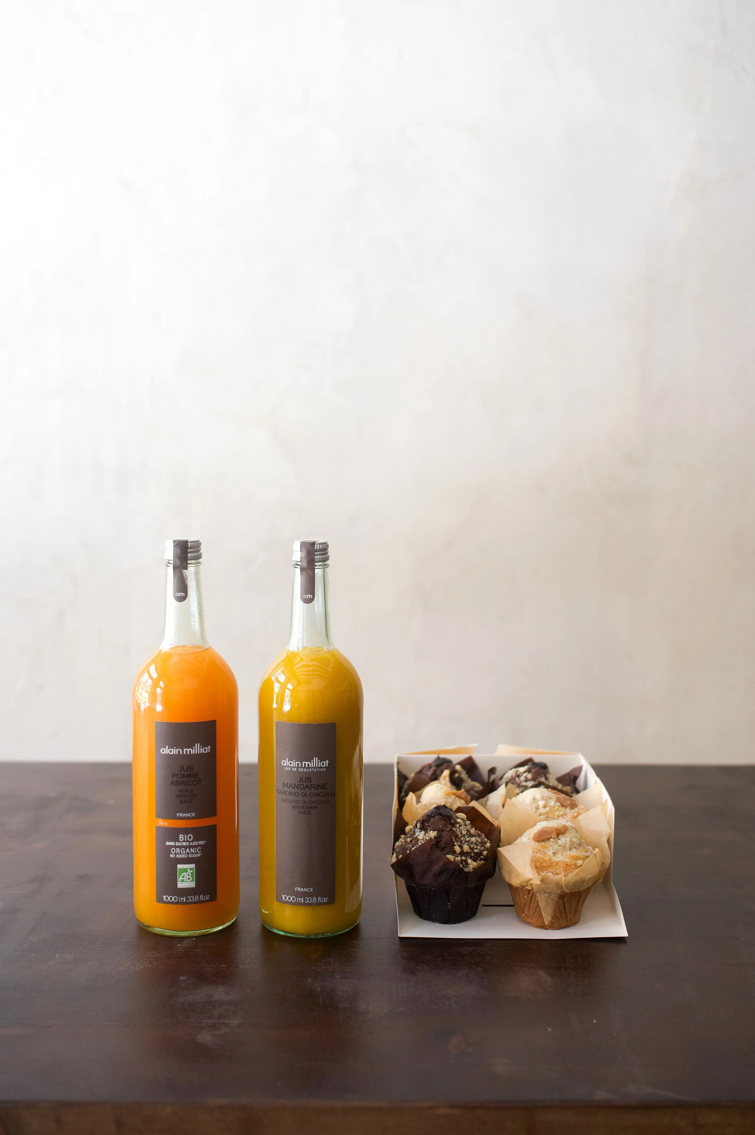 Deux bouteilles de jus de fruits, une à la pomme et à l'abricot, et l'autre à la mandarine, placées à côté d'un plateau de muffins aux chocolat et à la banane sur une table en bois.