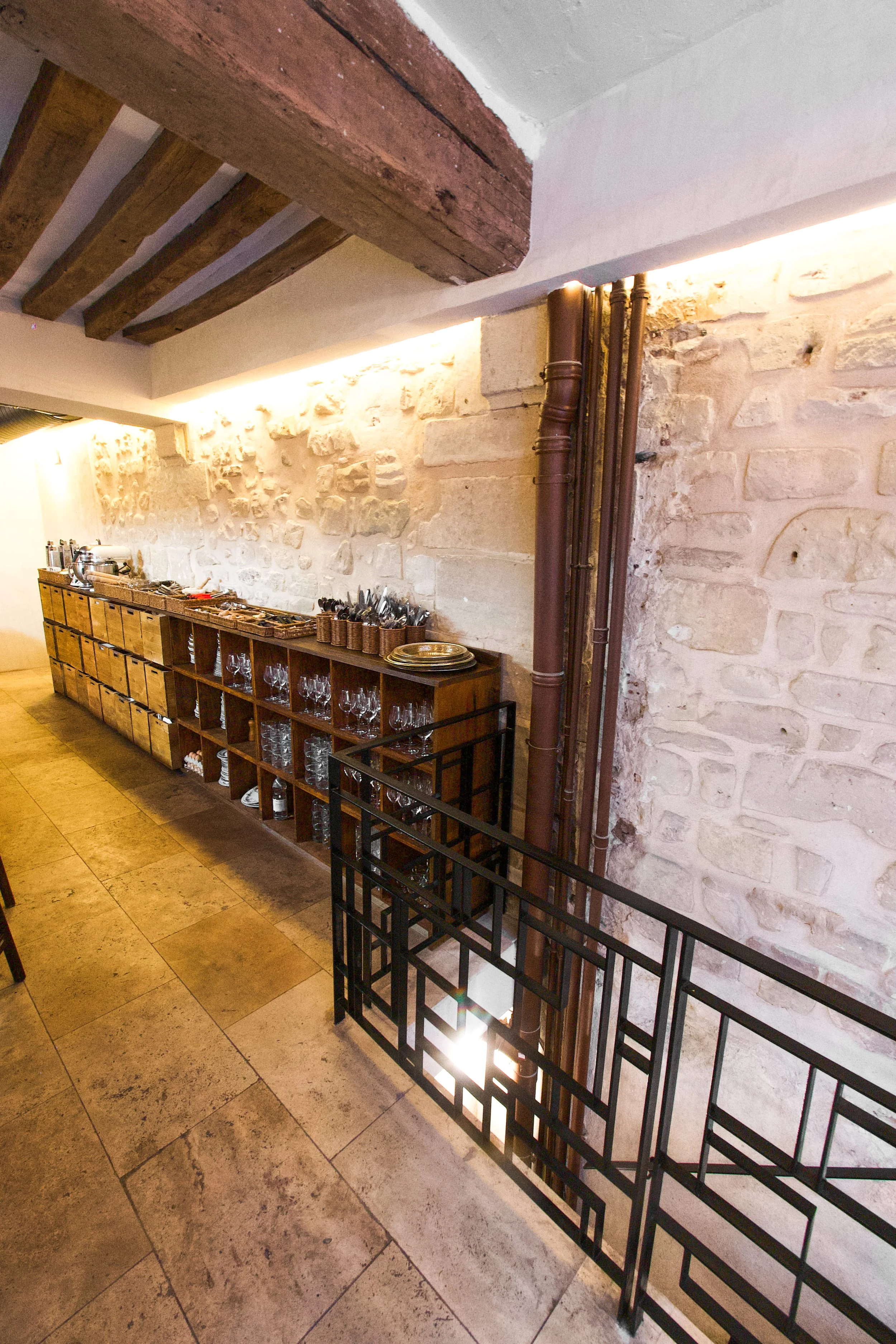 Dining area with wooden shelf containing glasses and plates, stone wall, wooden ceilings, warm lighting