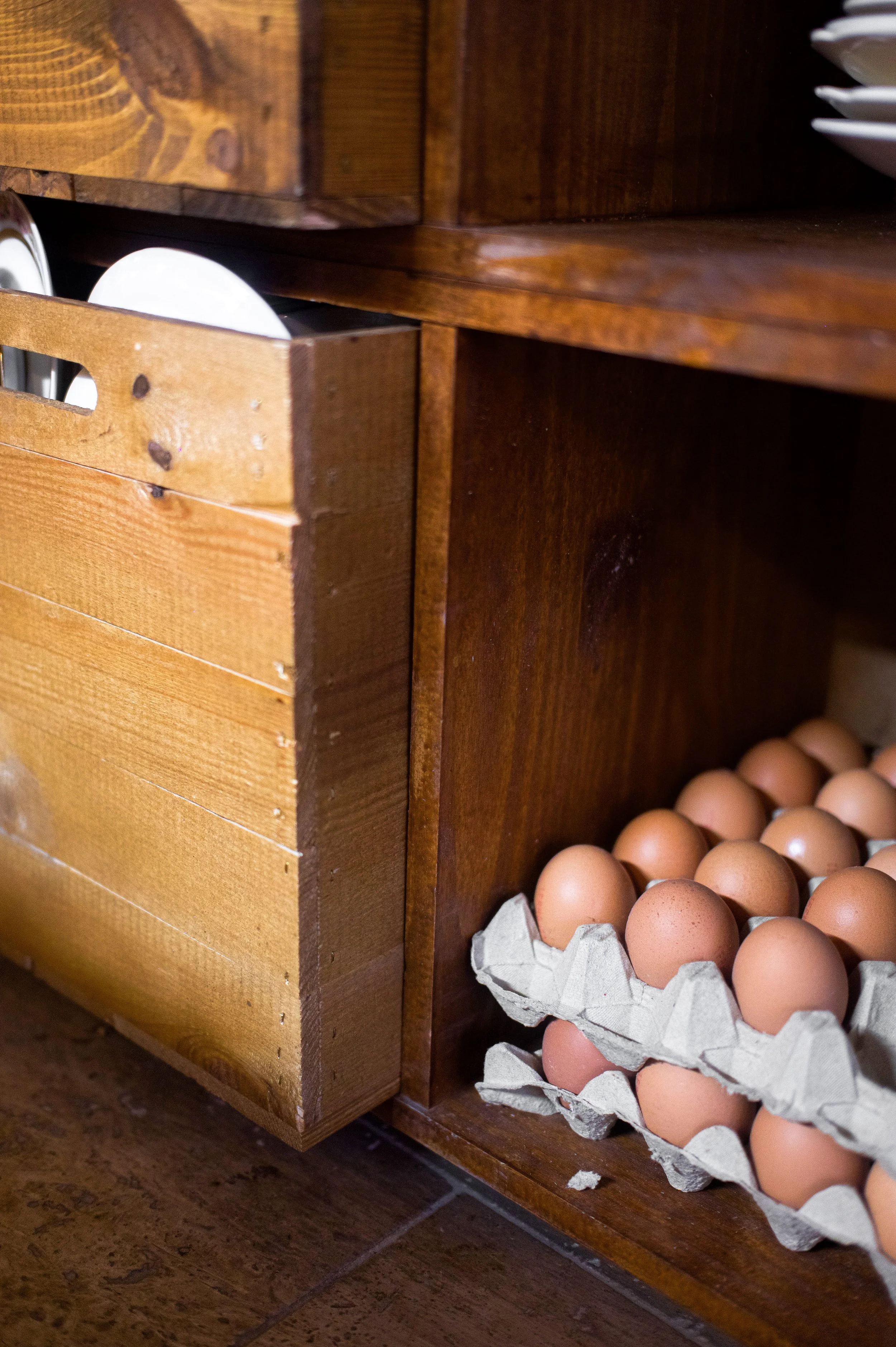 A wooden shelf containing a wooden box with plates, and a cardboard box filled with natural eggs in a kitchen.