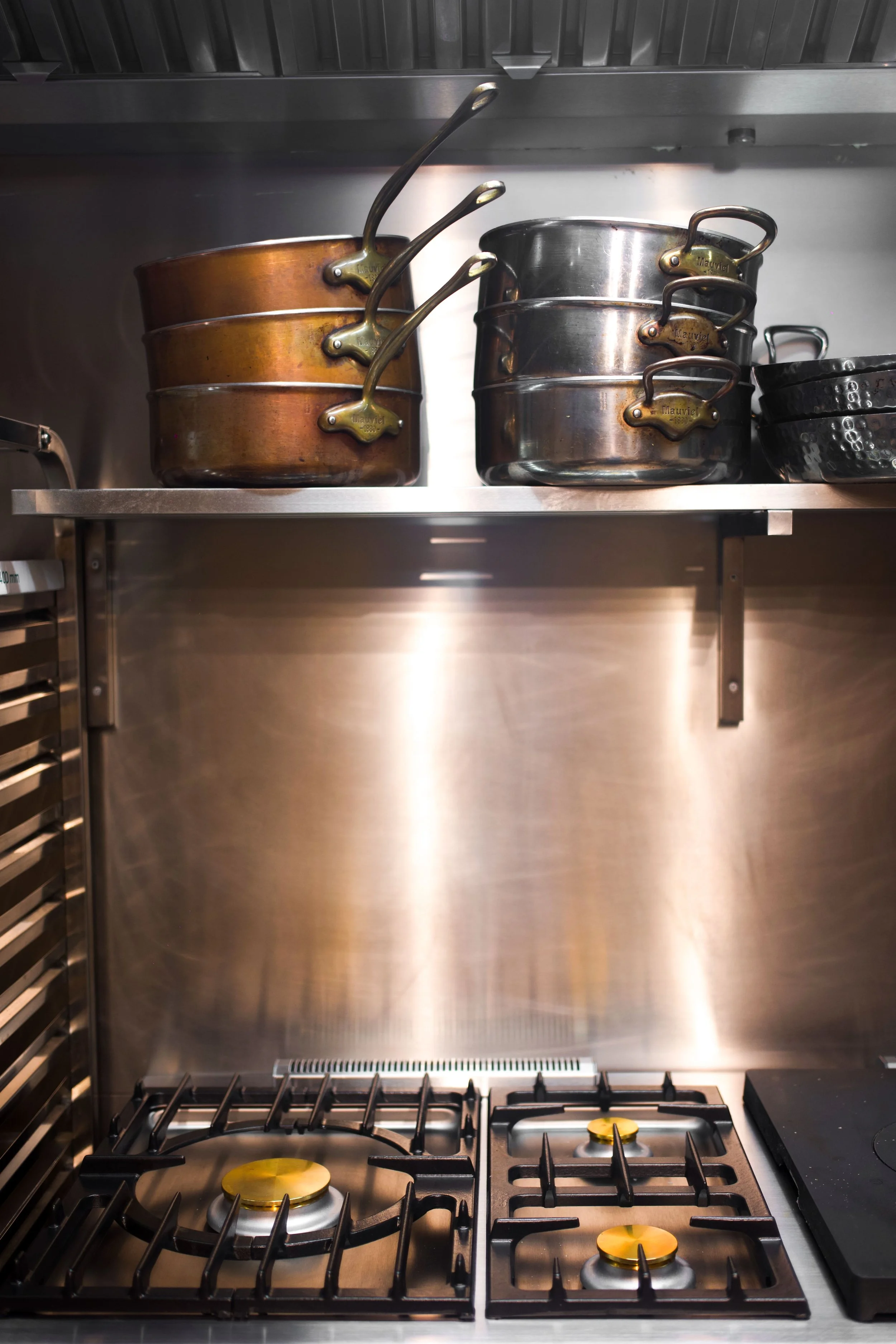 A stainless steel stove with two burners above and a four-burner hob below, with steel and copper pots arranged on a shelf above.