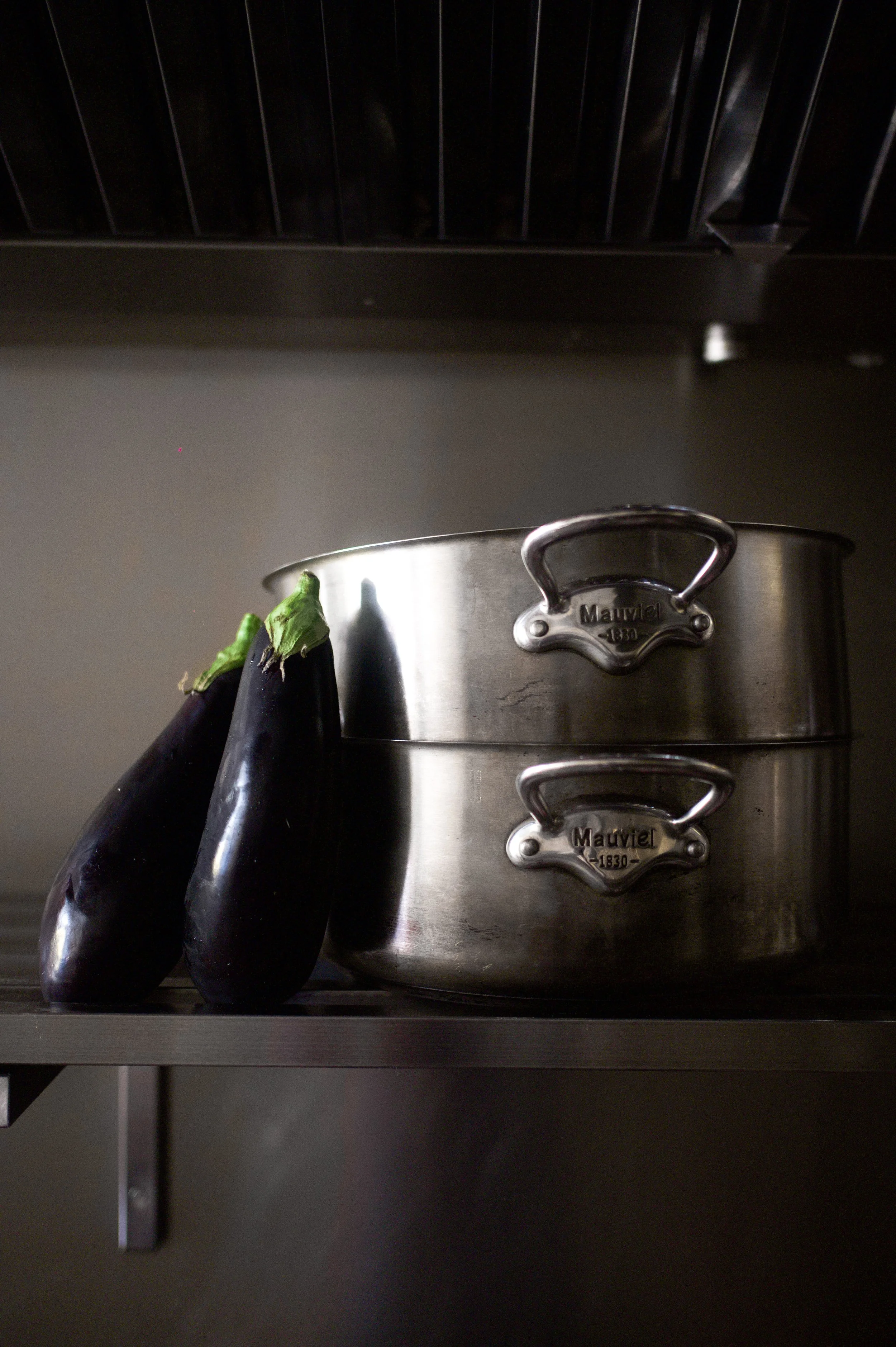 Deux aubergines noires posées côte à côte à côté d'une casserole en métal dans une cuisine.