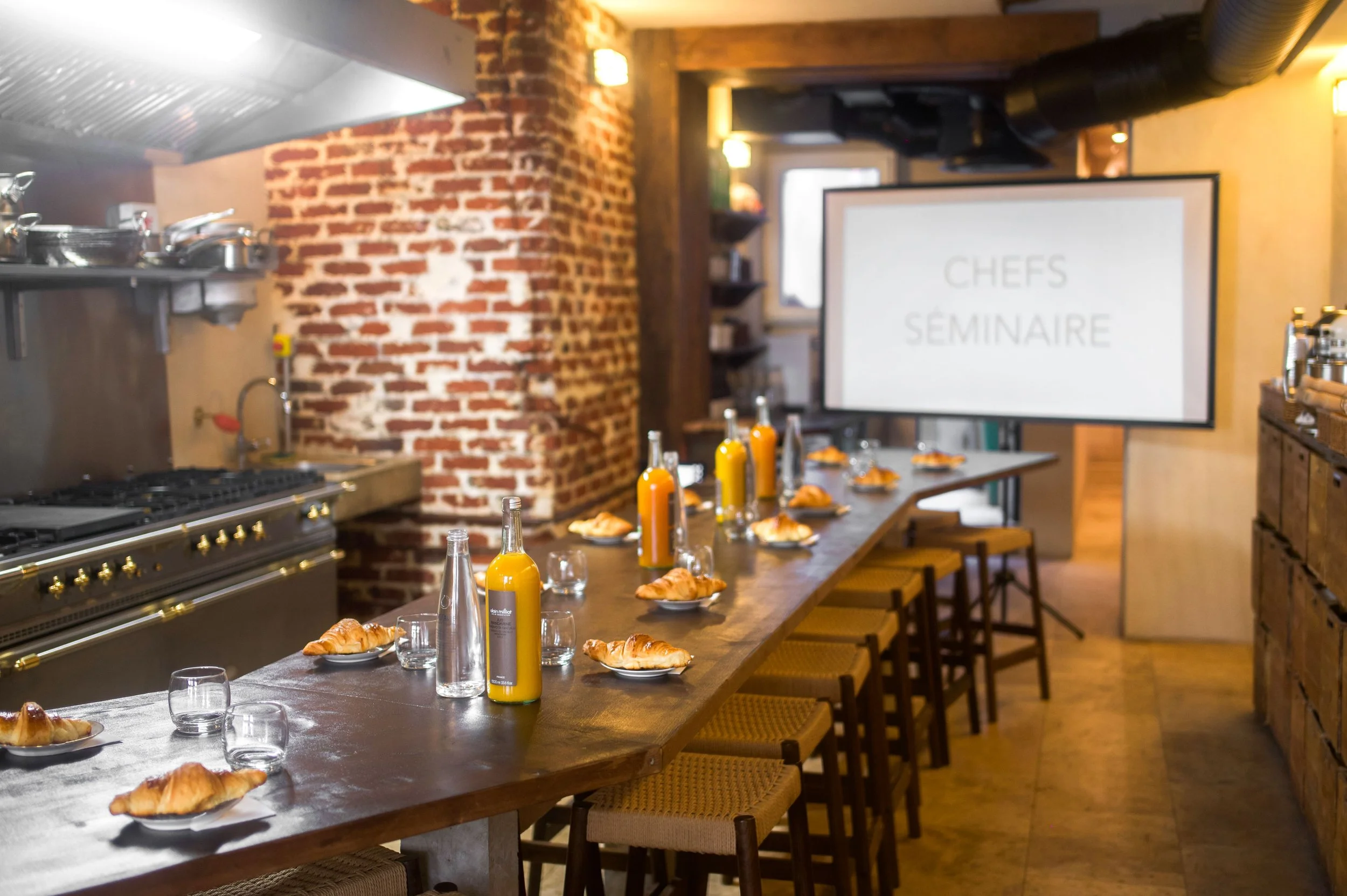 Salle de réunion avec une longue table en bois, croissants, bouteilles de jus, verres, écran affiche 'CHEFS SEMINAIRE', cuisine en arrière-plan, murs en briques et décor en bois.