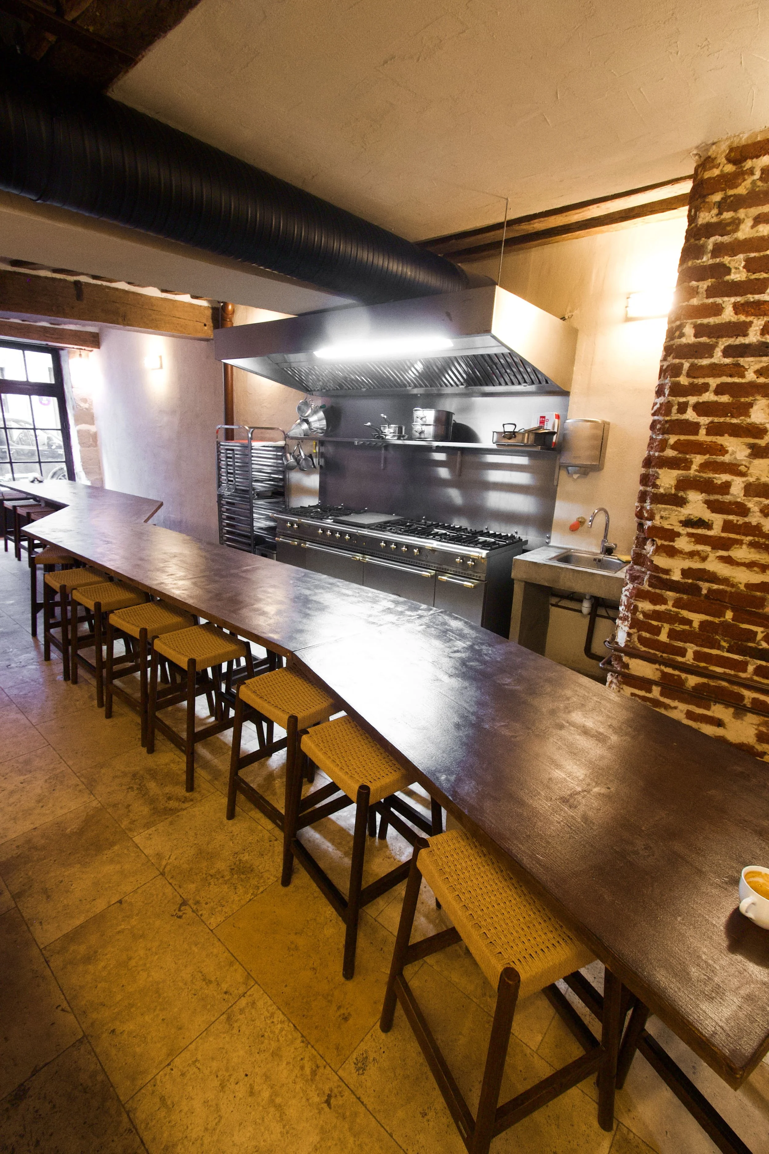 Restaurant kitchen with a large wooden table, wooden chairs with straw seats, and a professional kitchen in the background with hood, gas oven, pots, metal boards and sink.