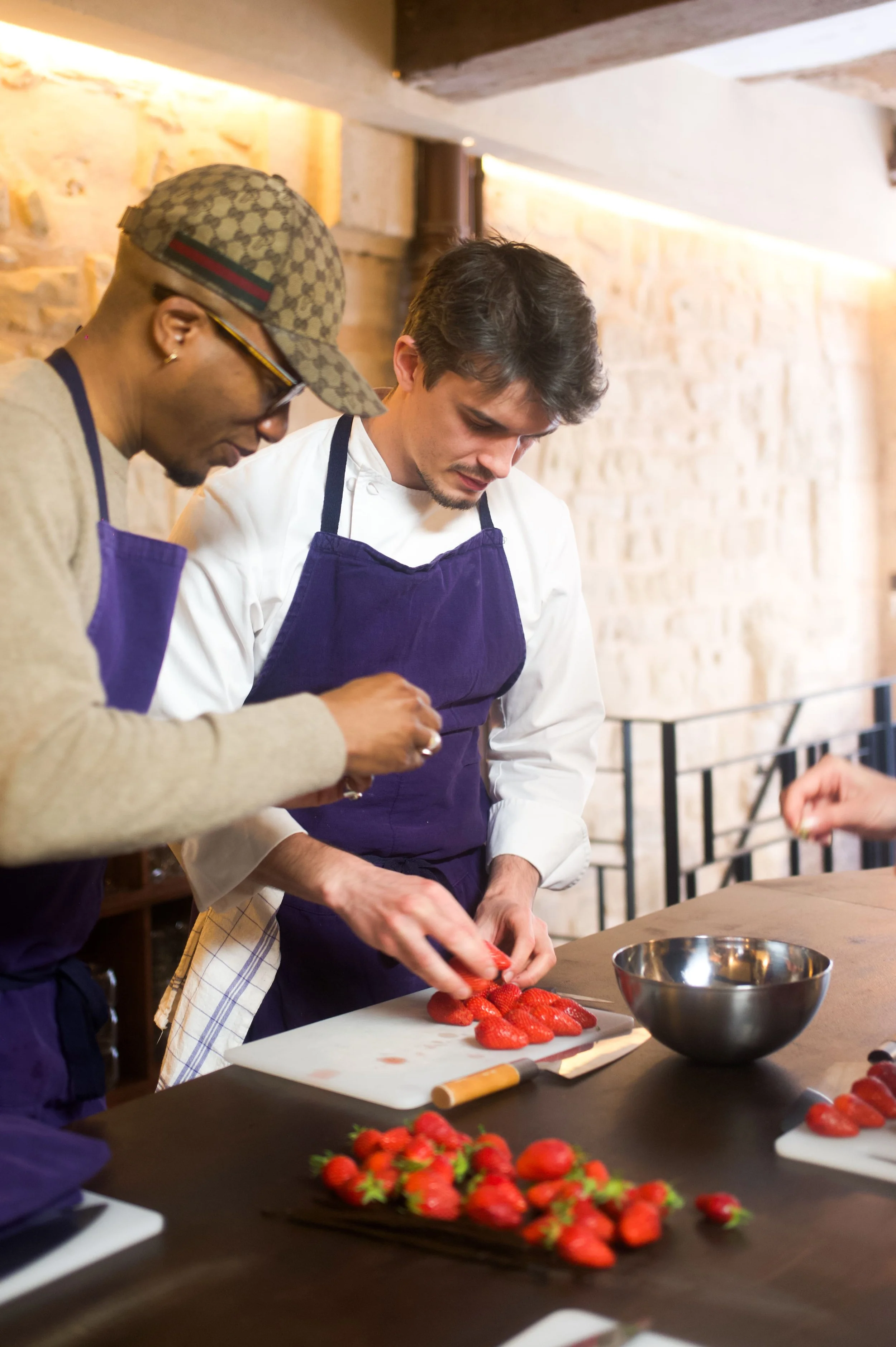 Deux hommes en tablier blanc et violet préparent des fraises dans une cuisine intérieure avec mur en pierre, utilisant une planche à découper.