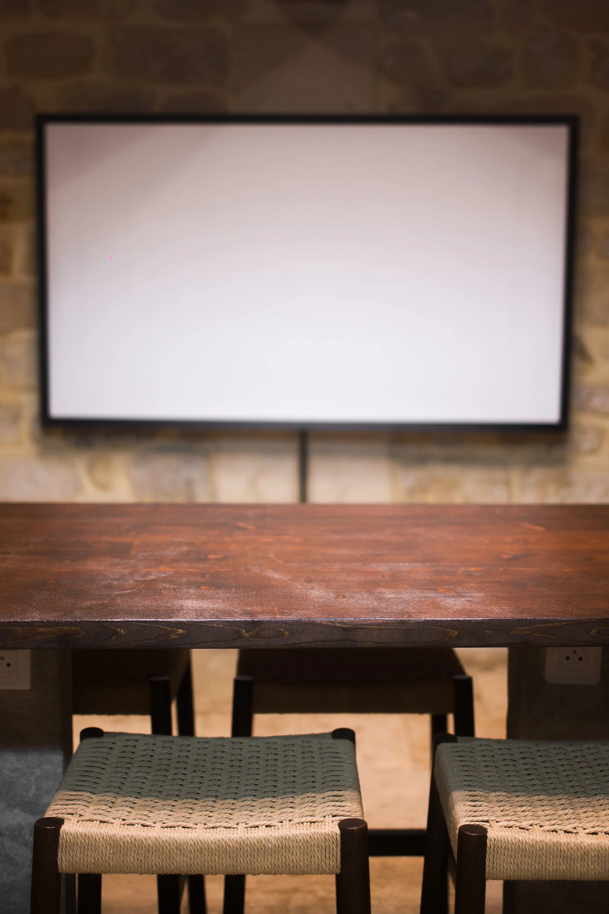 Dark wood table with two woven straw chairs, stone wall in background, white flat screen on wall