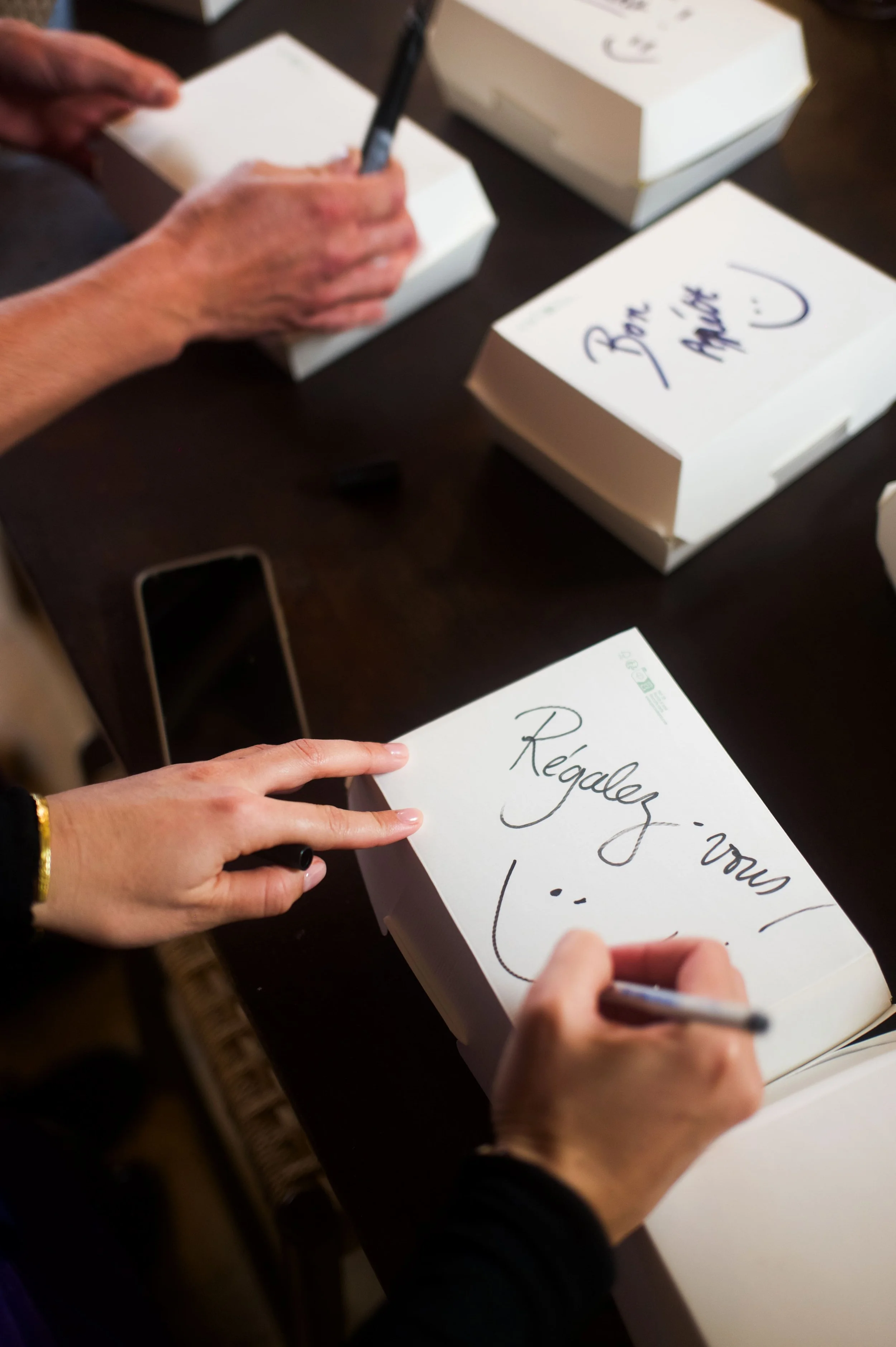 Une personne écrit à la main dans un livre blanc, avec d'autres livres similaires ouverts sur une table en bois. L'écriture dit « Regardez-nous ! » avec un smiley.