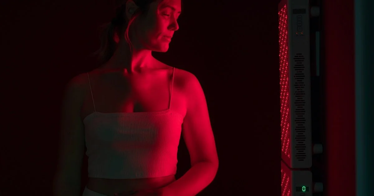 A woman in profile receives red light therapy from a tall, rectangular device emitting red light in a dark setting.