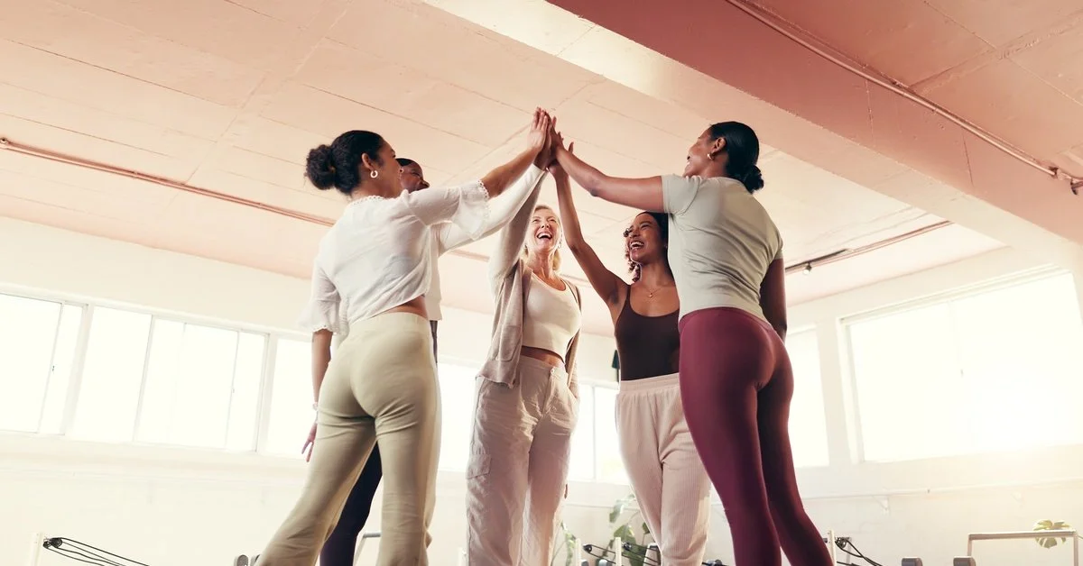 A low-angle view of 5 women giving each other high-fives as they stand in a circle in a brightly lit workout studio.