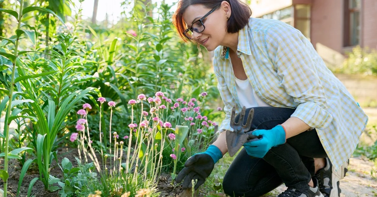 A woman in a plaid shirt and blue gloves kneels by an outdoor flower bed while holding gardening tools.