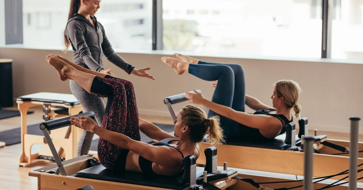 Two women perform Pilates on reformers while an instructor guides them in a bright studio with large windows.