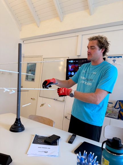 A man demonstrating a science experiment with a long, thin wire held between two supports, in a classroom or laboratory setting.