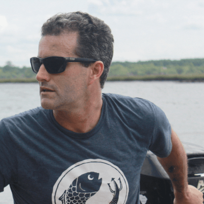 Man wearing sunglasses and a navy T-shirt with a fish design, standing outdoors near water.