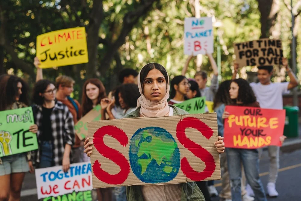 A young woman with a beige headscarf holding a handmade sign featuring a drawing of the Earth with the words "SOS" in red around it, at a climate protest with other protesters holding signs advocating for climate justice and environmental action.