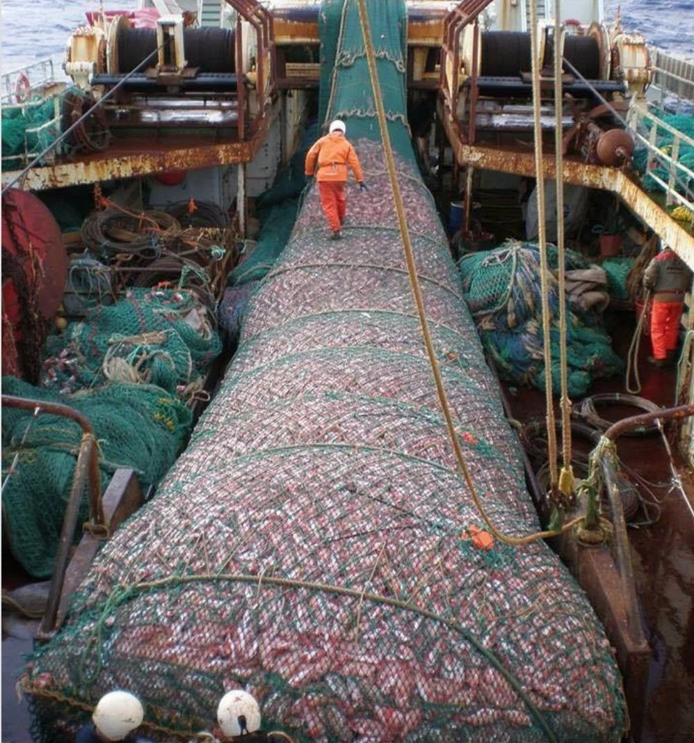 A fishing boat with a large net on deck, two crew members in orange jackets, large spools of rope, and various fishing gear.