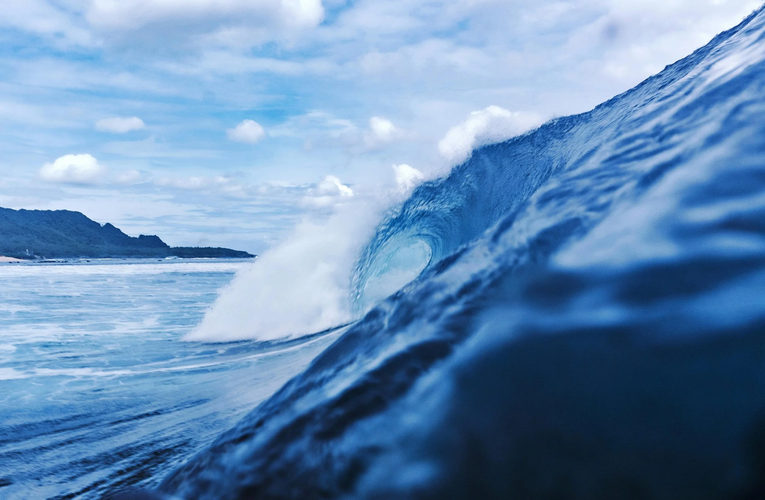 Ocean wave curling with a distant shoreline and cloudy sky in the background.