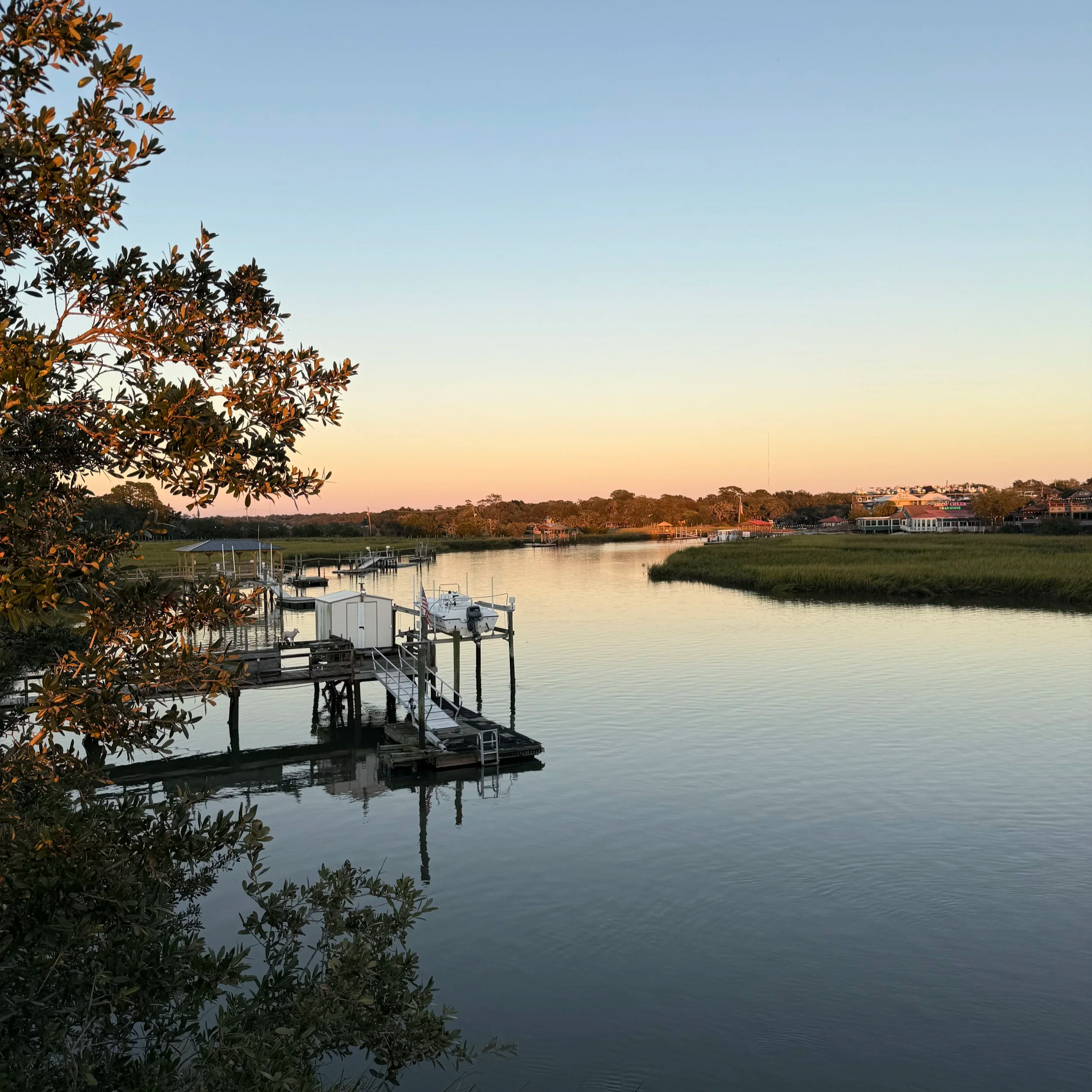 Calm river scene at sunset with a dock and boat, surrounded by greenery and houses in the background.