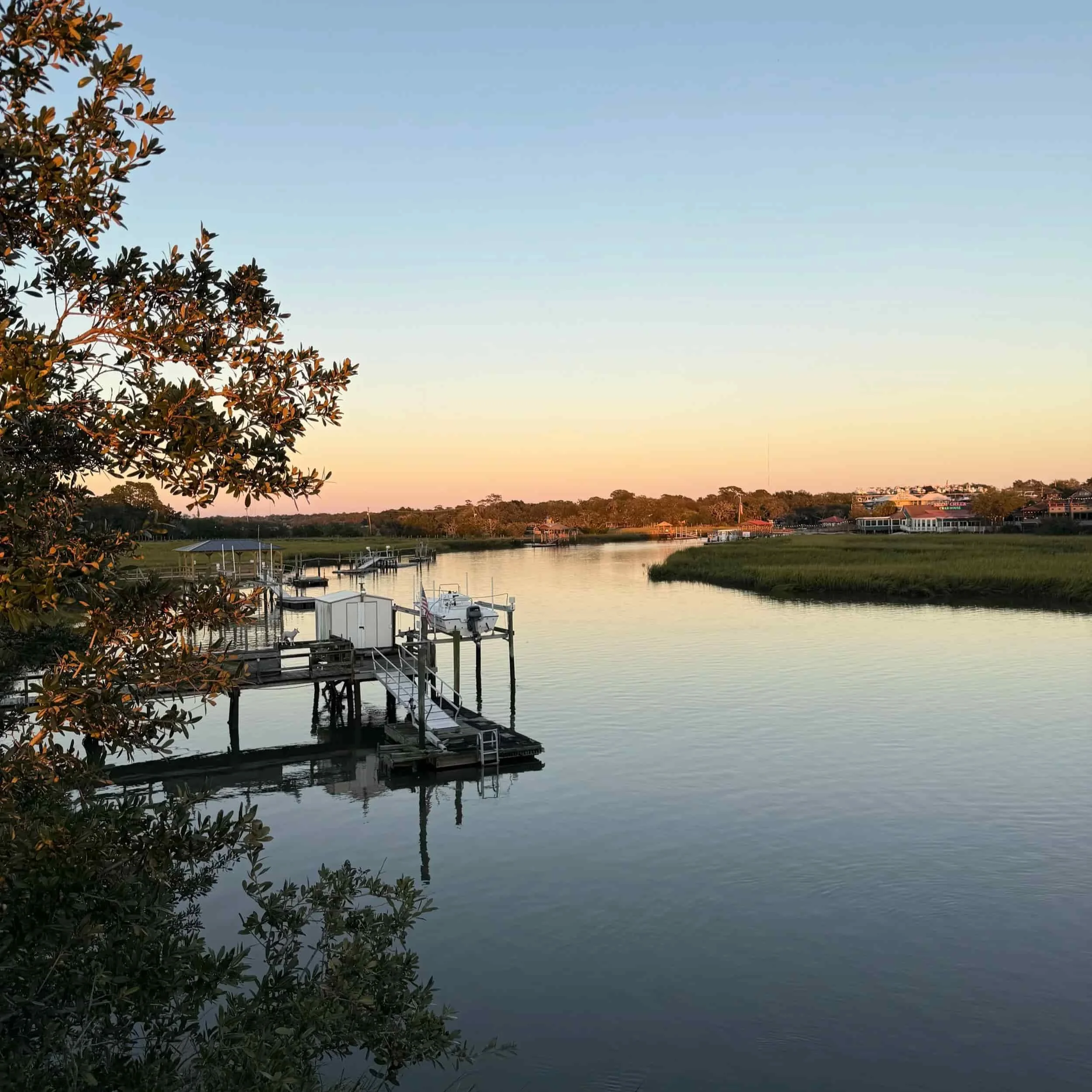 A calm waterway at sunset with docks and boats and houses in the distance, framed by trees.