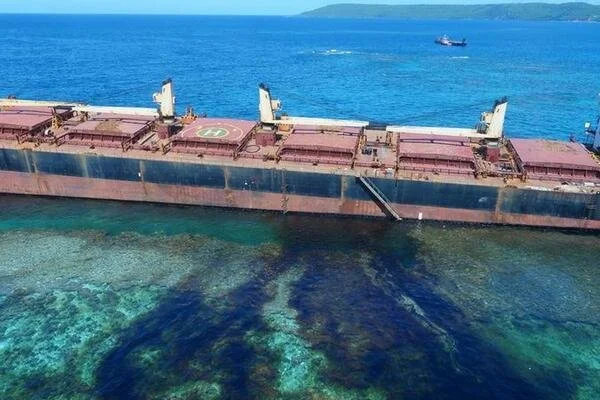 A large, partially submerged shipwreck lying in shallow, clear blue water near a coastline.
