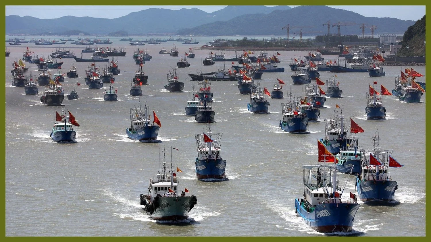 A large number of fishing boats with red flags on a body of water, possibly during a parade or celebration, with hills and cranes in the background.