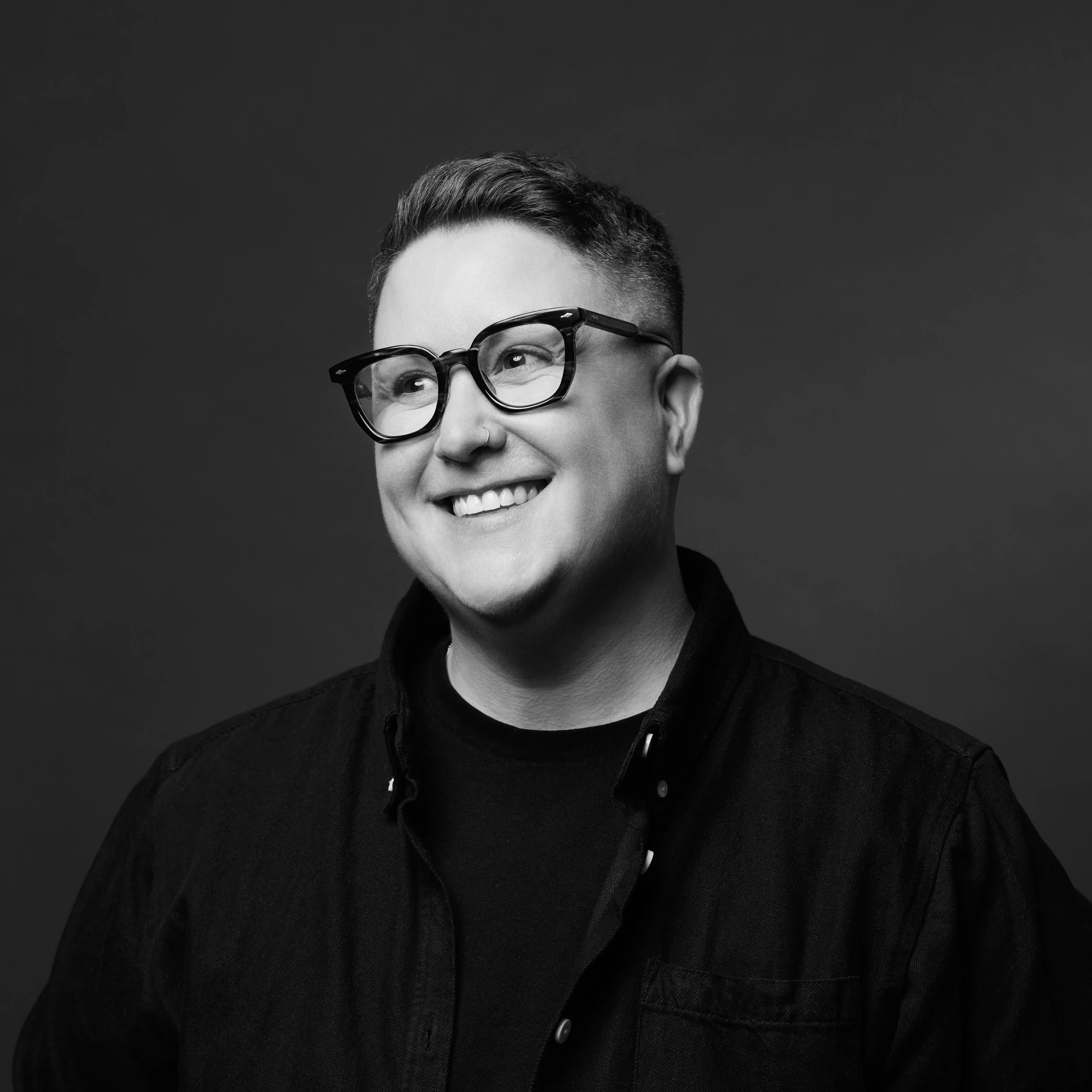 Close-up black and white portrait of a person with short hair, smiling, wearing large, dark-rimmed glasses, and a dark jacket against a dark background.