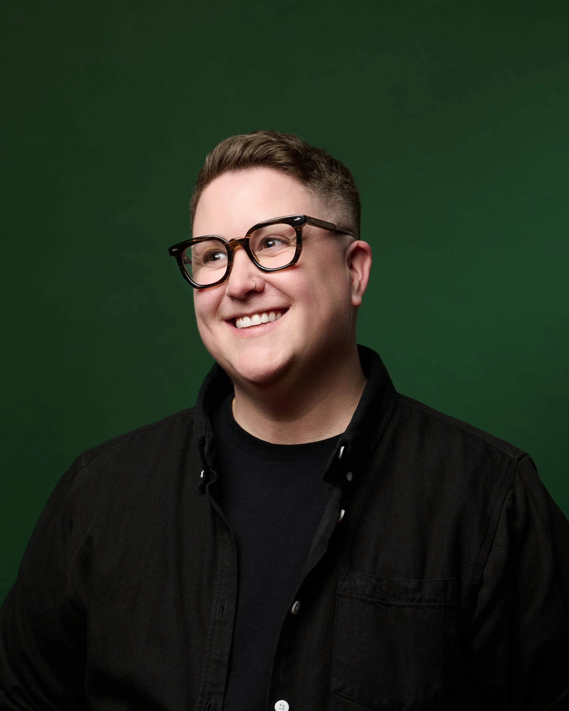 A full colour image of a white nonbinary person in a black shirt wearing glasses on a green background smiling looking off to the left