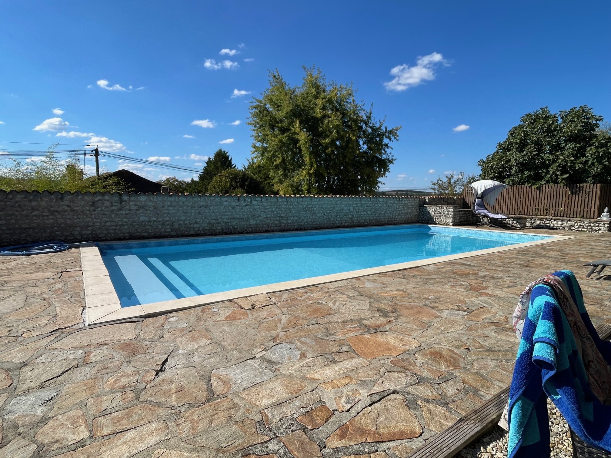 Outdoor swimming pool surrounded by stone patio, with lush trees and a clear blue sky in the background.