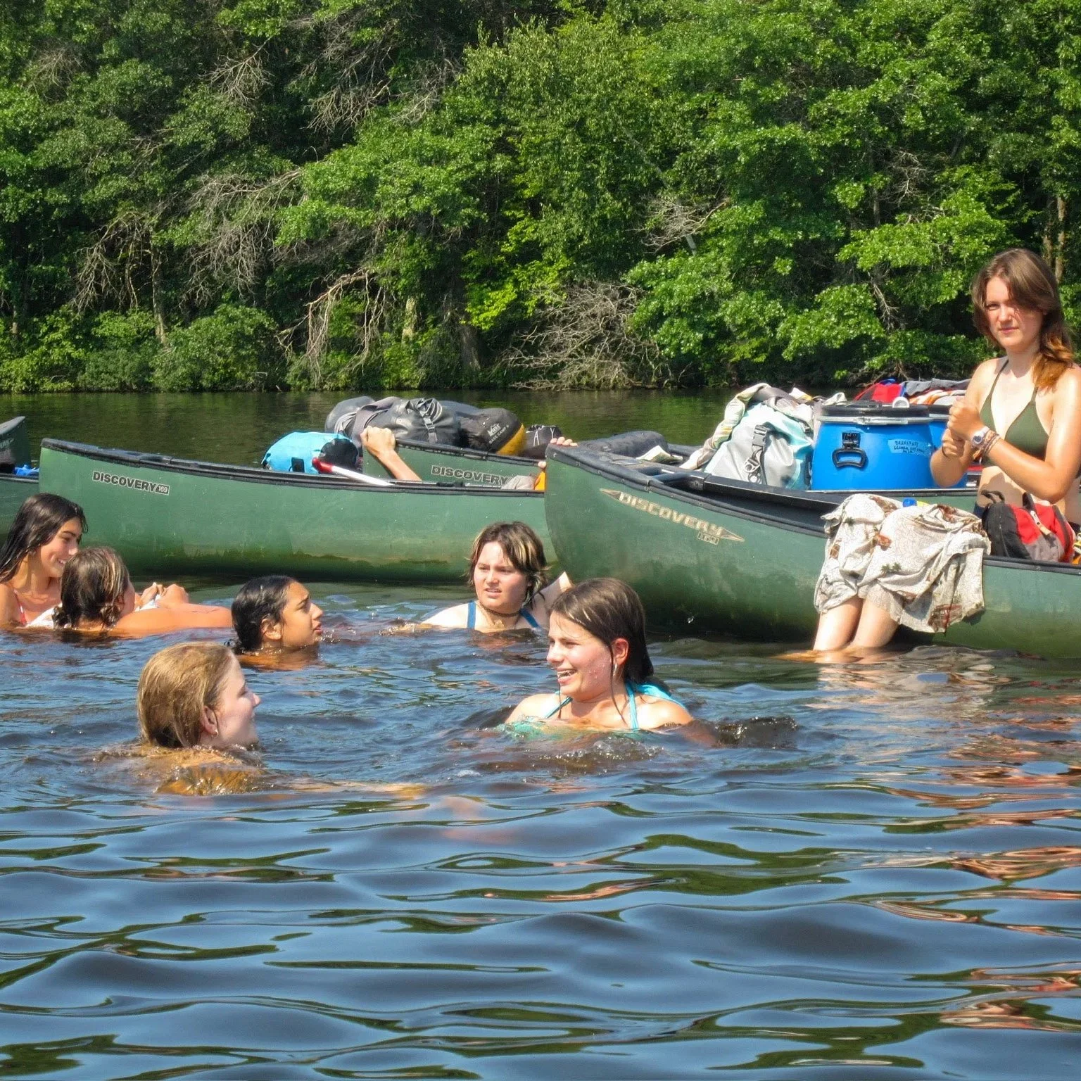 Group of people swimming and relaxing in a lake with two green canoes in the background, surrounded by trees.