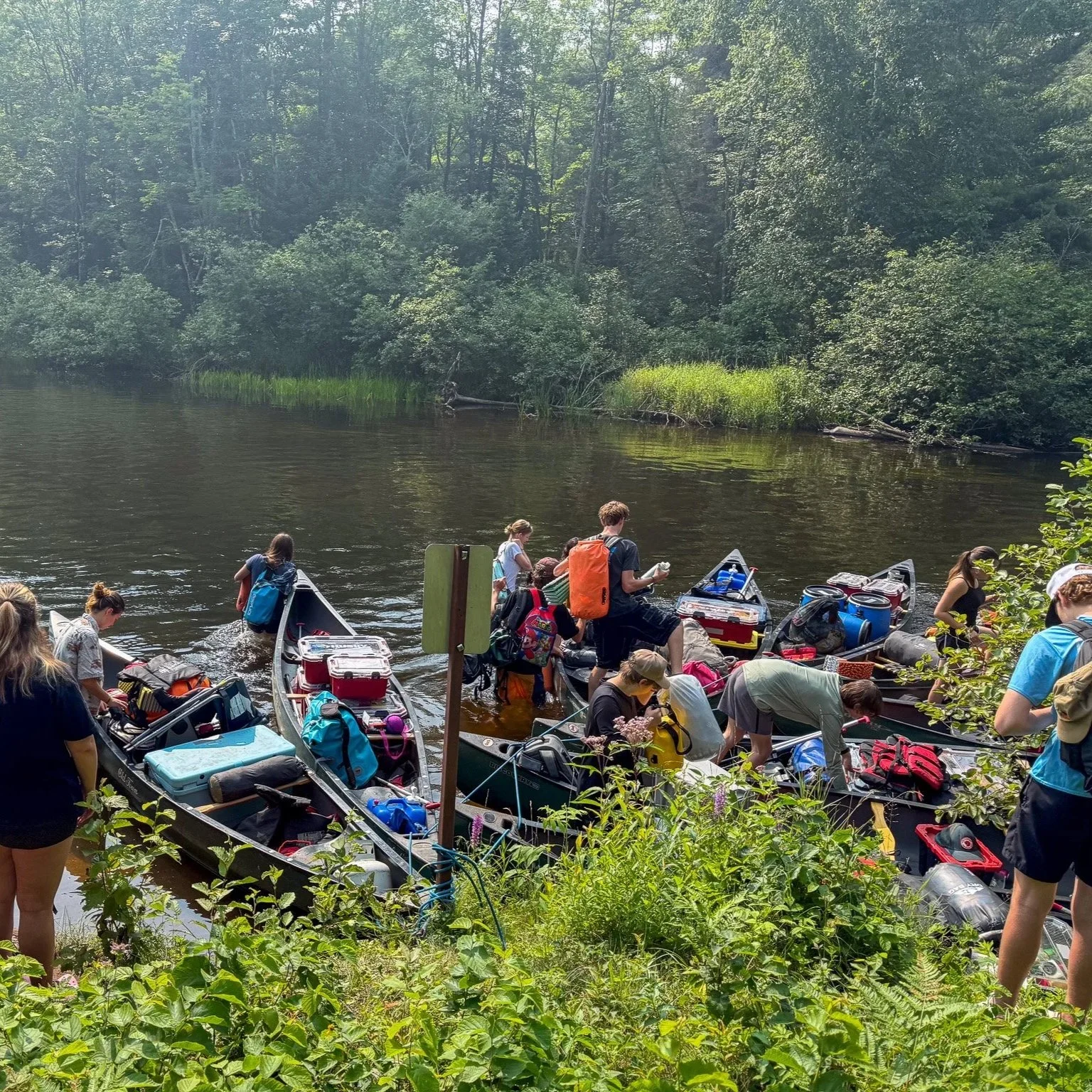 Group of people preparing to launch canoes into a river surrounded by lush green trees and bushes.