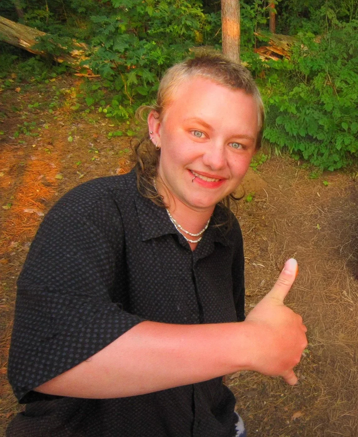 A woman with light skin, short blond hair, blue eyes, earrings, and a lip piercing, wearing a black textured shirt, smiling, and giving a thumbs up in an outdoor wooded area.
