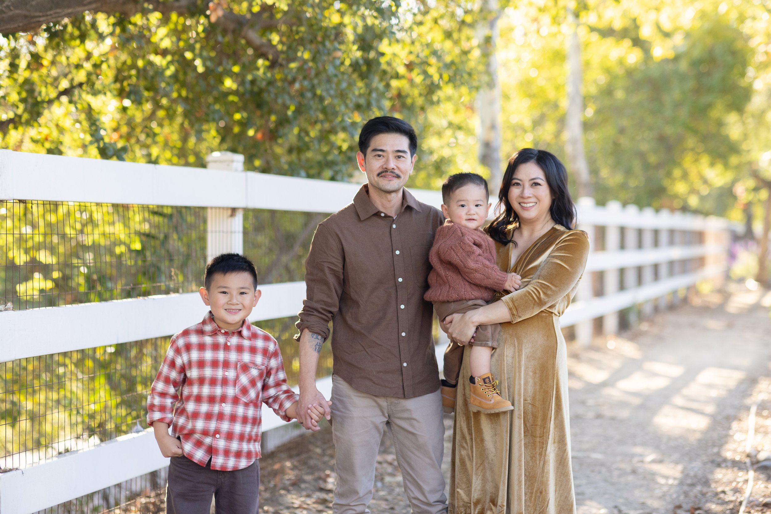 Family of five standing on a dirt path outdoors during autumn, smiling at the camera, with a white fence and trees with yellow leaves in the background.