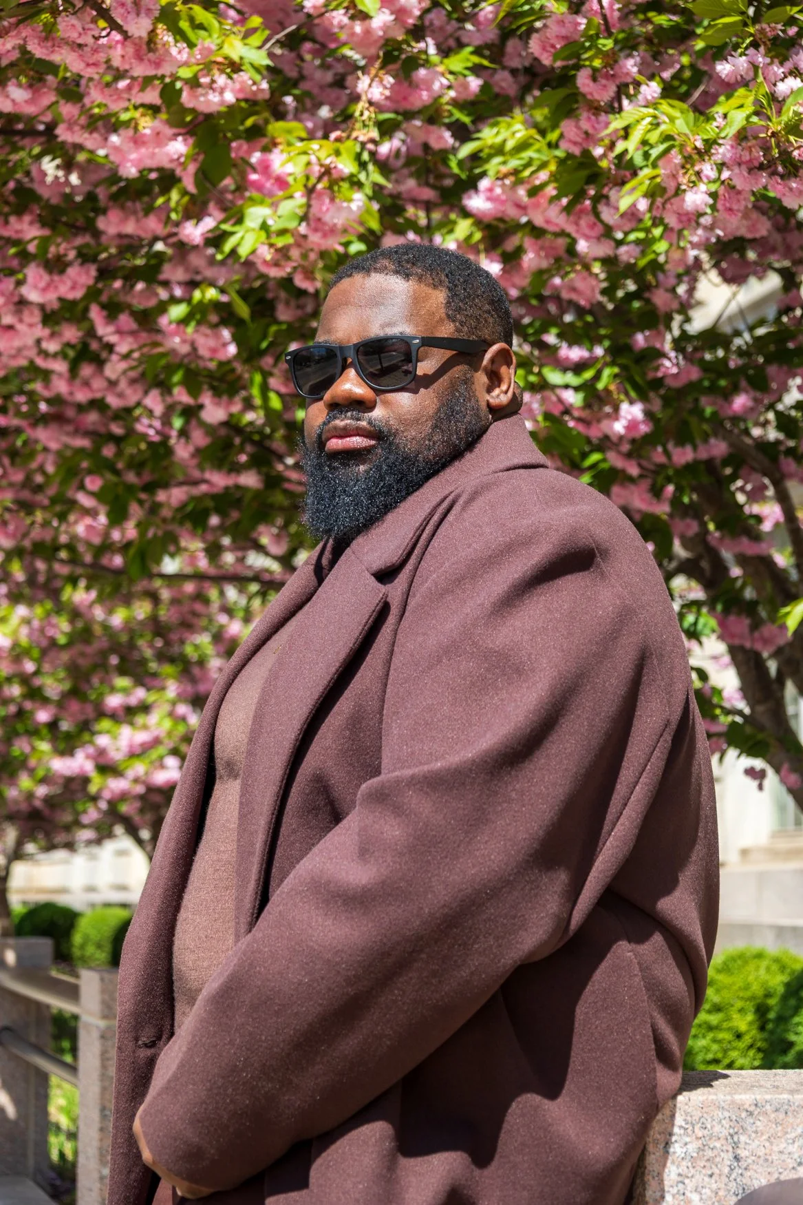 Outdoor portrait of a man wearing a burgundy jacket and sunglasses posing in front of flowering trees.