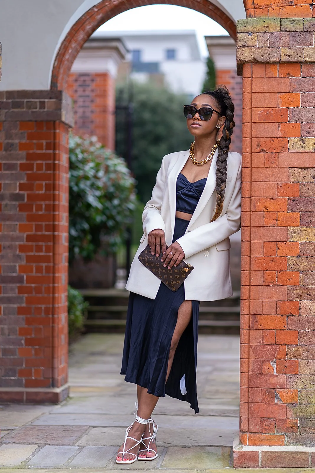 Fashionable woman with long braid wearing sunglasses, white blazer, navy dress with slit, holding a designer clutch, standing under an archway.