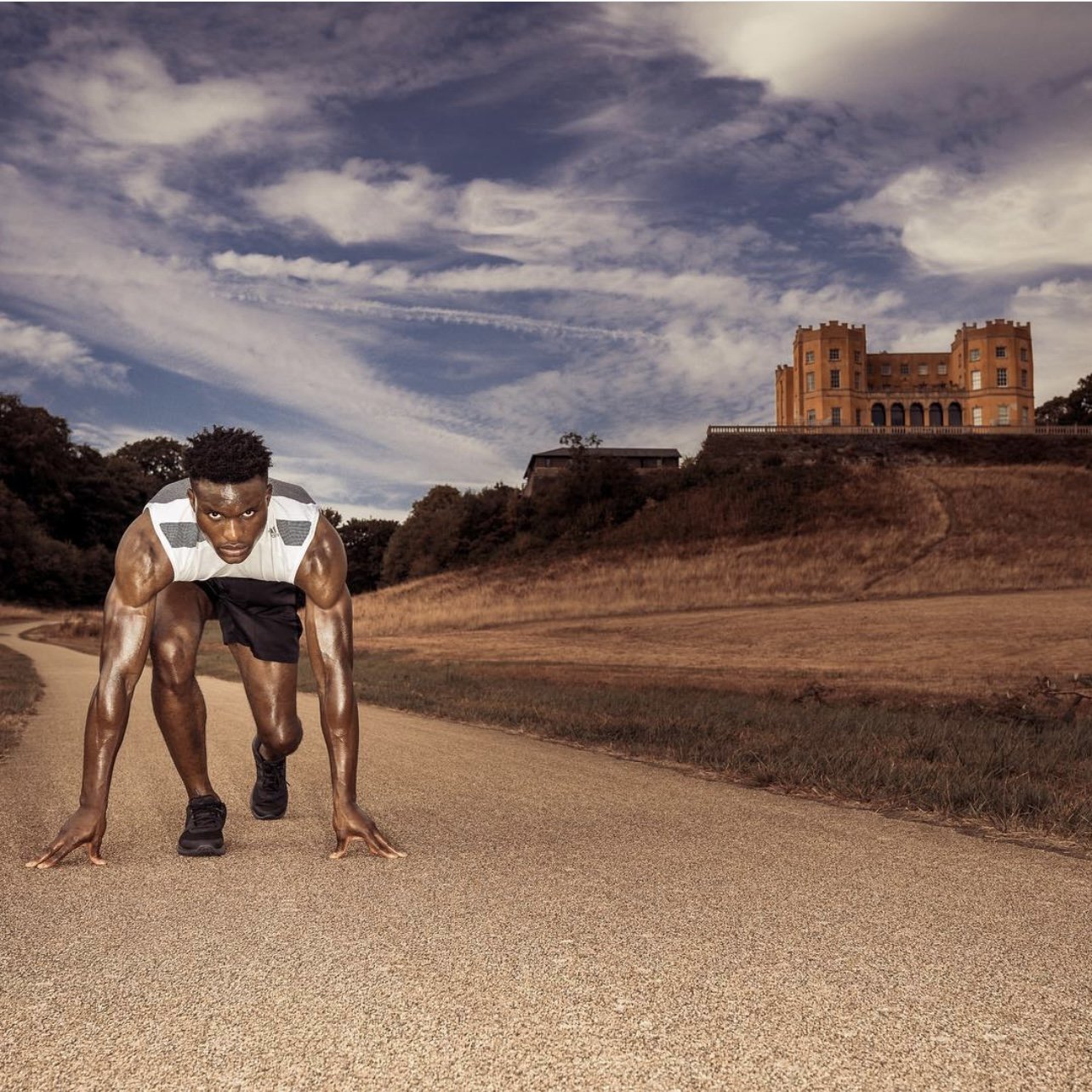 Athlete in starting position on country road with castle in background, cloudy sky.