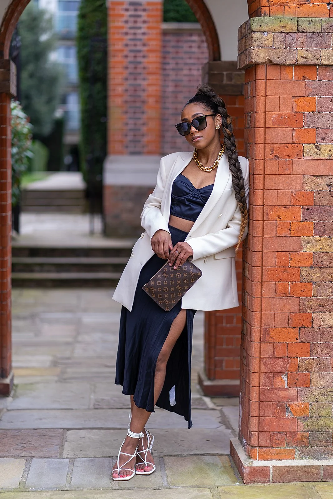Fashionable woman in a stylish outfit with long braid, white blazer, black top, black skirt, sunglasses, gold chain necklace, holding designer clutch, posing against brick archway.
