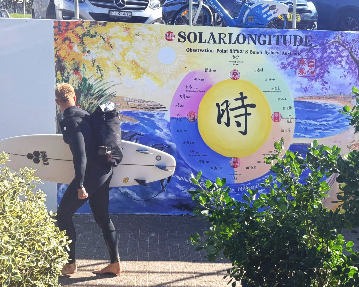 A surfer carrying a board past Solar Longitude – Time Orbit at Bondi Sea Wall, connecting the public artwork with Bondi’s coastal life.