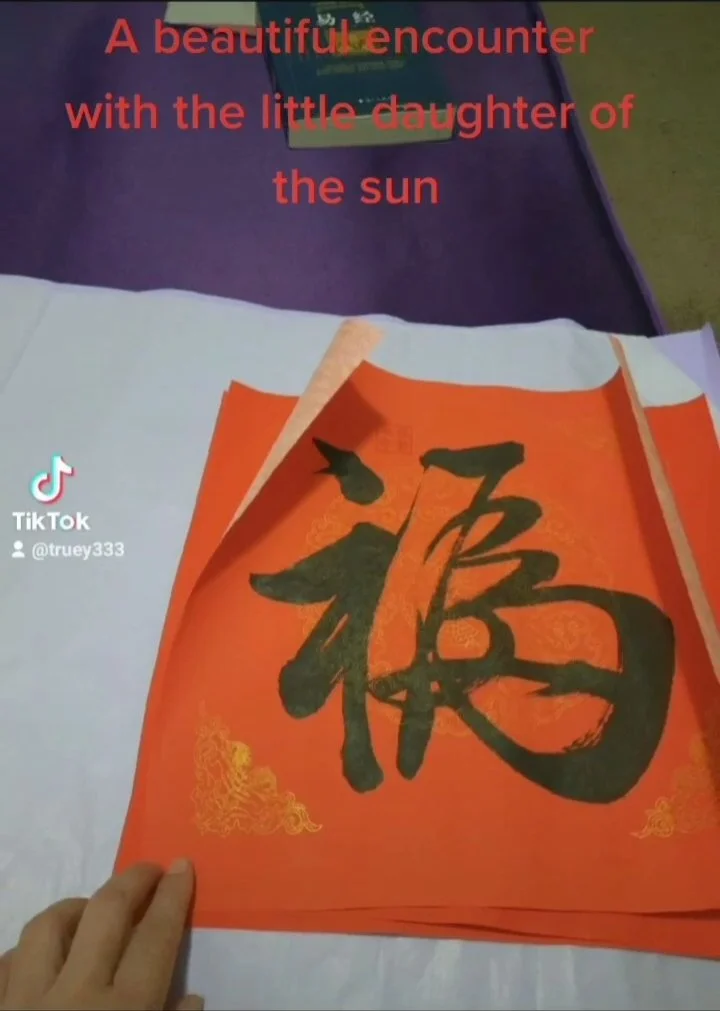Colorful Chinese calligraphy art with large black character on red paper, on a white surface. Overlaid red text says, "A beautiful encounter with the little daughter of the sun."